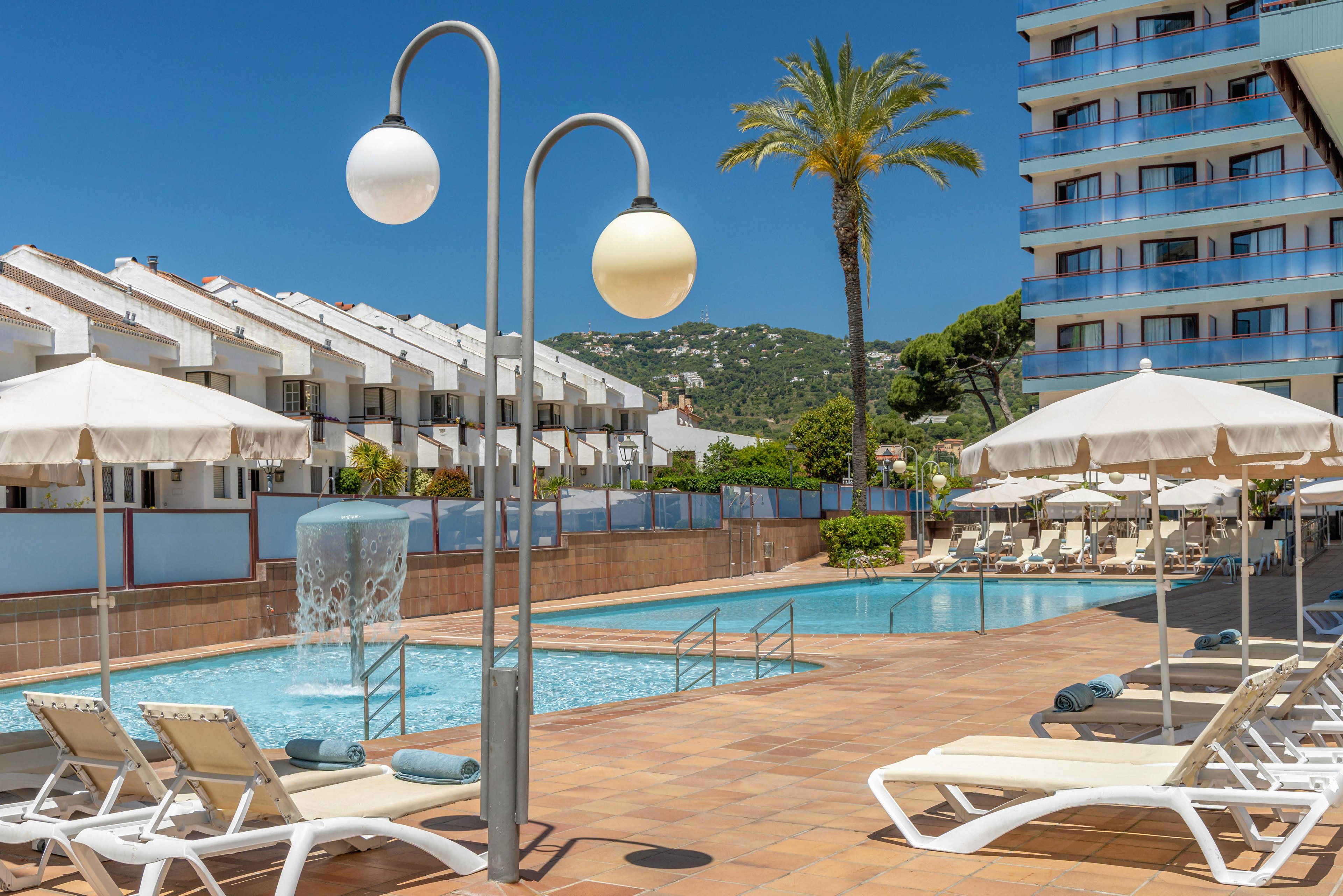 An outdoor resort swimming pool is surrounded by white sun loungers and umbrellas on a bright sunny day. A modern streetlamp and a small waterfall feature stand near the pool steps. In the background, white townhouses and a taller hotel building rise against green hills. A single palm tree and clear blue sky enhance the relaxed vacation atmosphere.