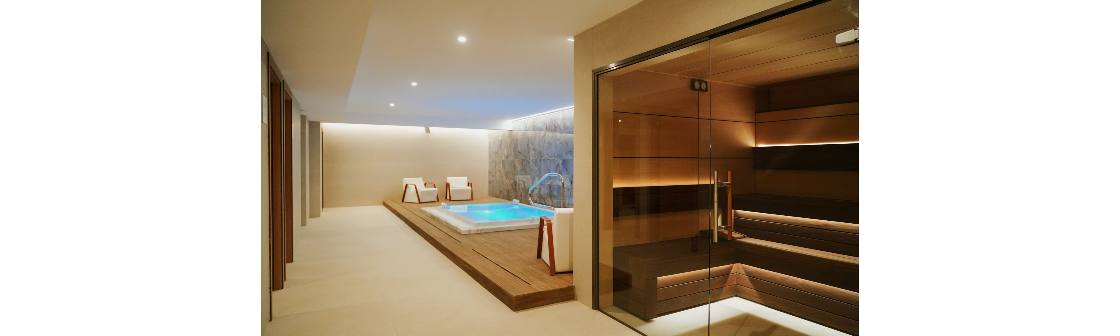 A contemporary spa interior features a glowing jacuzzi set into a wooden platform. Two white lounge chairs sit nearby against a stone accent wall, creating a relaxing retreat. On the right, a warm wooden sauna is enclosed by a large glass wall and door. Soft recessed lighting and neutral tones enhance the calm, luxurious atmosphere.