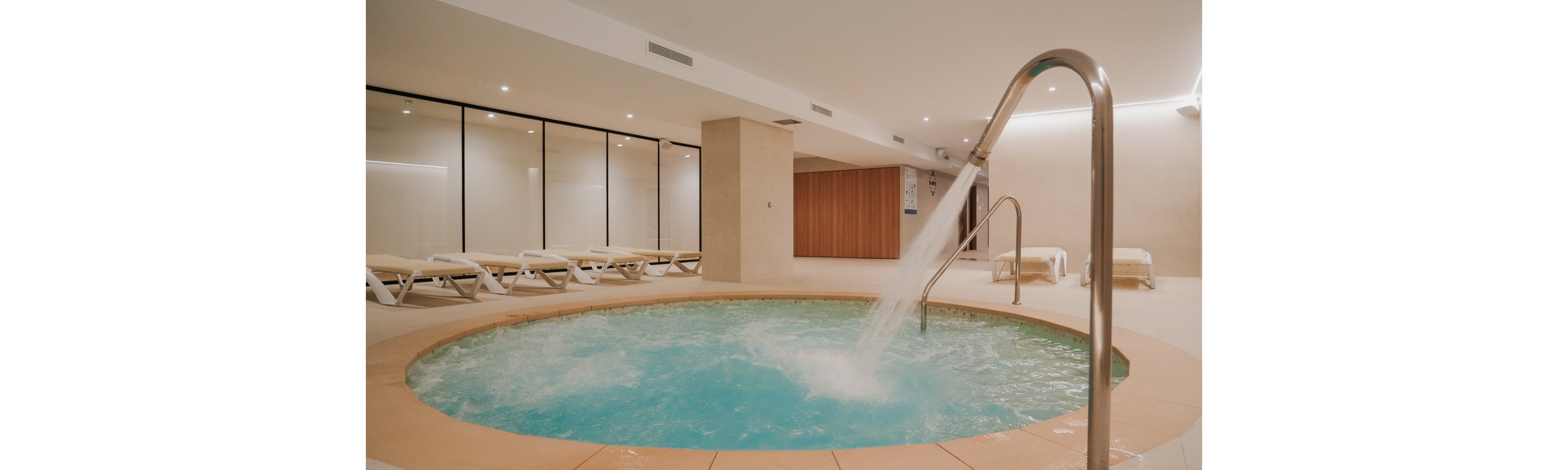 Circular indoor jacuzzi pool with bubbling water sits in a modern spa room. A curved metal fountain pipe pours a strong stream into the turquoise water. Several white lounge chairs line the wall behind glass partitions, suggesting a relaxation area. Warm neutral walls and soft ceiling lighting create a calm, minimalist atmosphere.