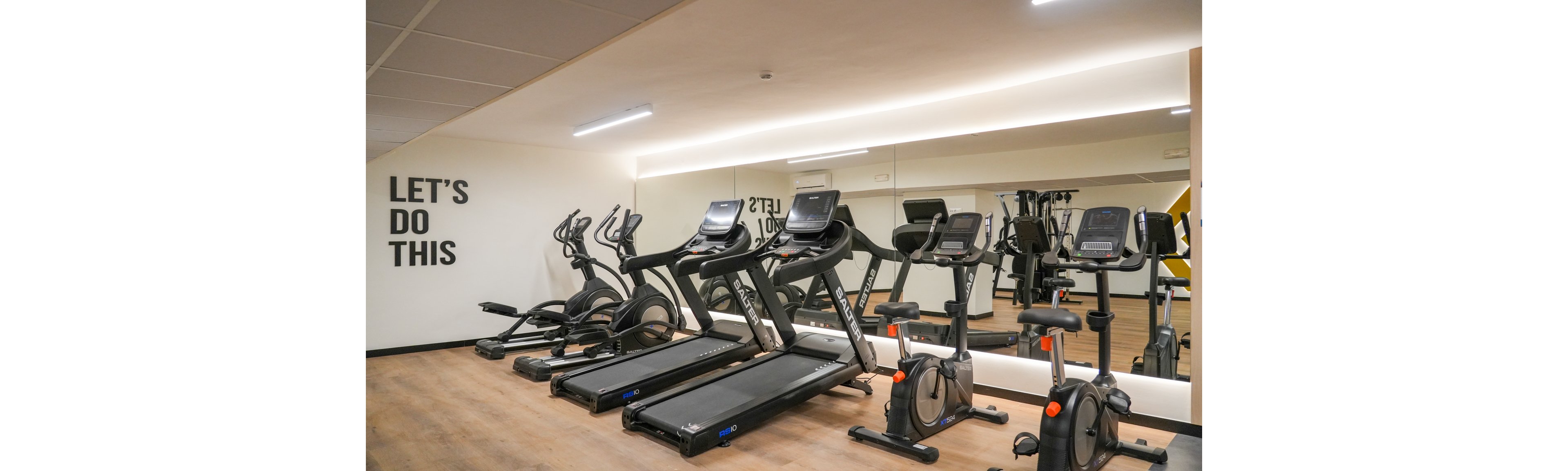 A row of treadmills and stationary exercise bikes is arranged neatly in a modern indoor gym. The equipment faces a large wall mirror that reflects the machines and bright ceiling lights. Wooden flooring and clean white walls create a minimalist, professional fitness environment. Motivational text on the wall reads "LET'S DO THIS," encouraging an energetic workout atmosphere.