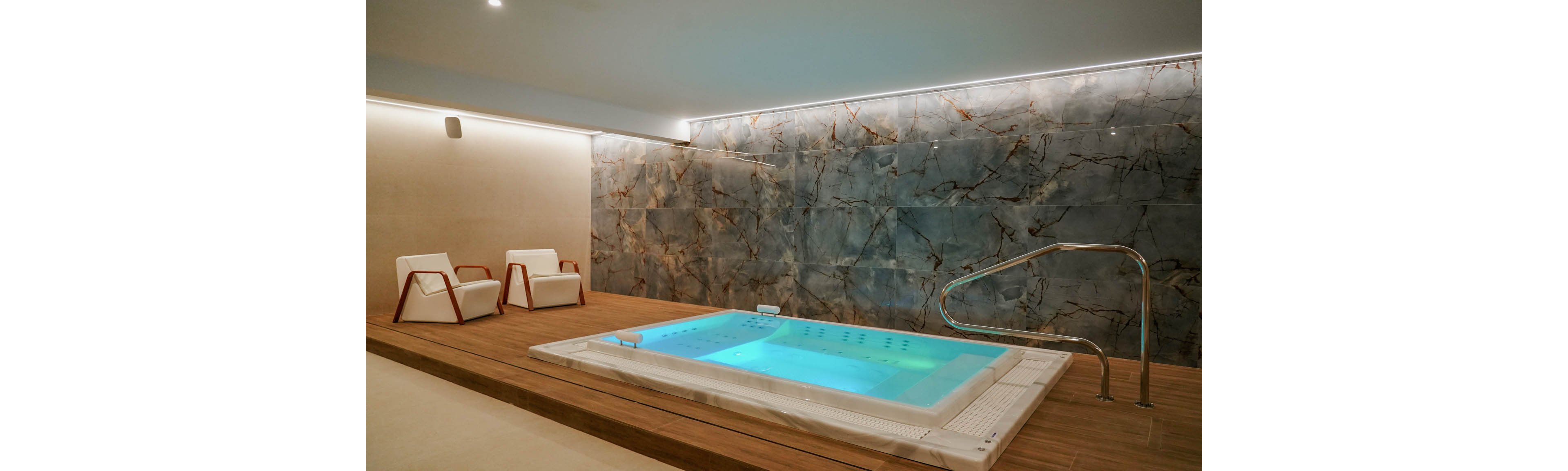 A modern indoor spa room features a rectangular jacuzzi pool set into a wooden platform. Soft turquoise lighting illuminates the bubbling water, contrasting with the warm tones of the wood. Two contemporary white lounge chairs sit nearby for relaxation. A large stone-textured wall and subtle ceiling lights create a calm, luxurious atmosphere.