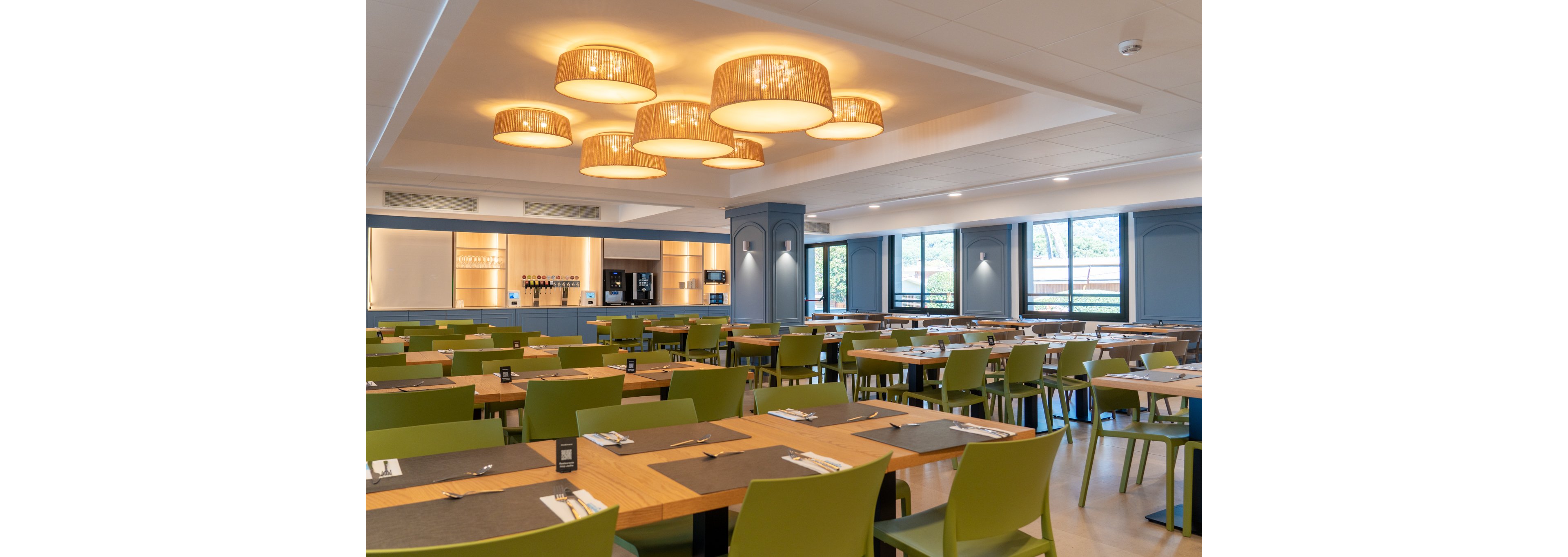 The image shows a spacious cafeteria with neatly arranged wooden tables and green chairs. The setting is bright and contemporary, featuring large windows and stylish ceiling lights with woven shades. Place settings with cutlery and placemats are visible on each table, and a beverage station is seen in the background. The overall mood is inviting and clean, with a neutral palette accented by green and wood tones.