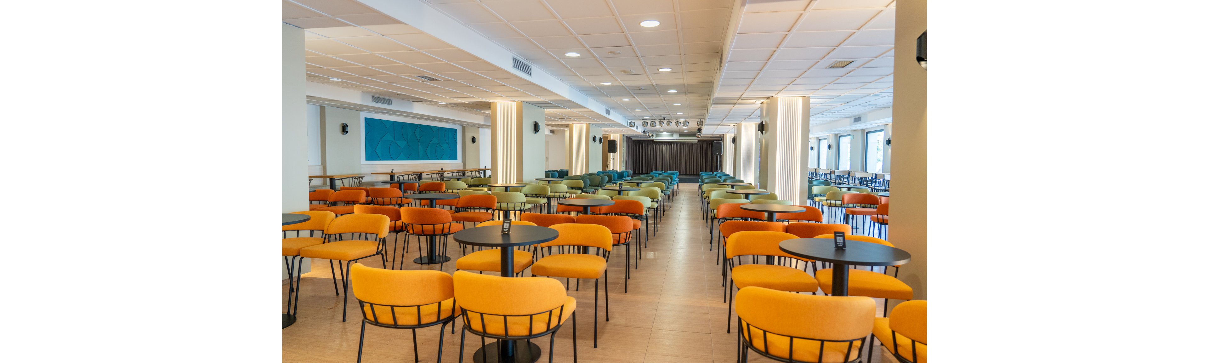 A spacious cafeteria features rows of round tables paired with vibrant orange and green cushioned chairs. The setting is well-lit with ceiling lights and large windows, creating a bright and inviting atmosphere. The room is empty, emphasizing the clean, organized layout and contemporary design elements.