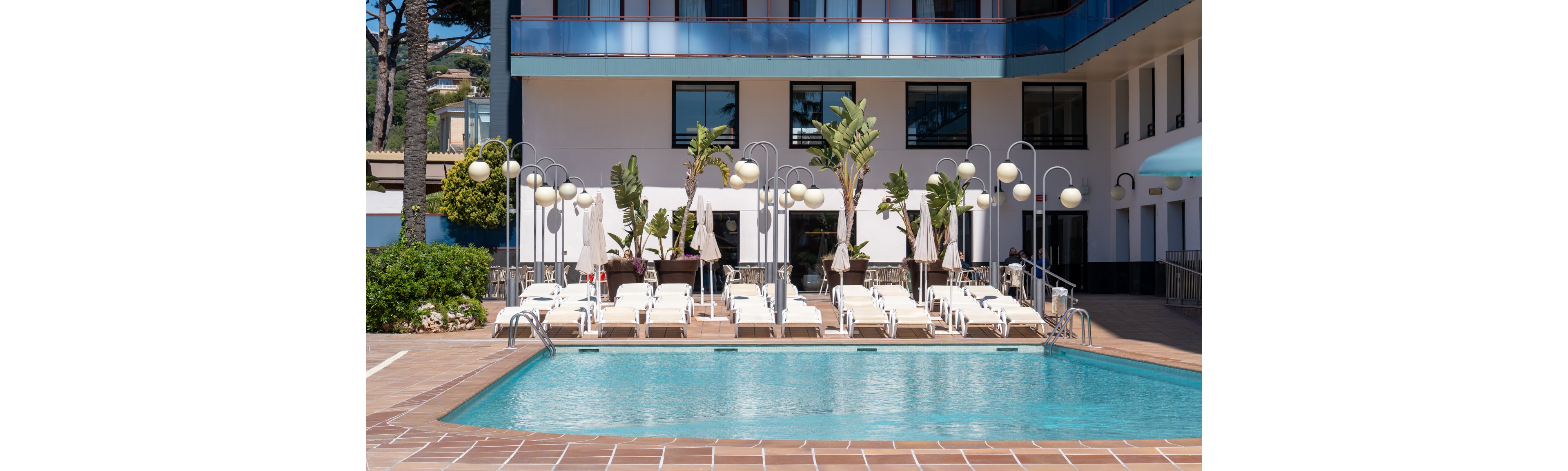 The image features a contemporary hotel swimming pool surrounded by neatly arranged white sun loungers and umbrellas. Tall palm trees and decorative lamp posts add a tropical touch to the poolside area. The hotel building has large windows and a clean, minimalist facade. The setting is bright and sunny, evoking a relaxed vacation mood.