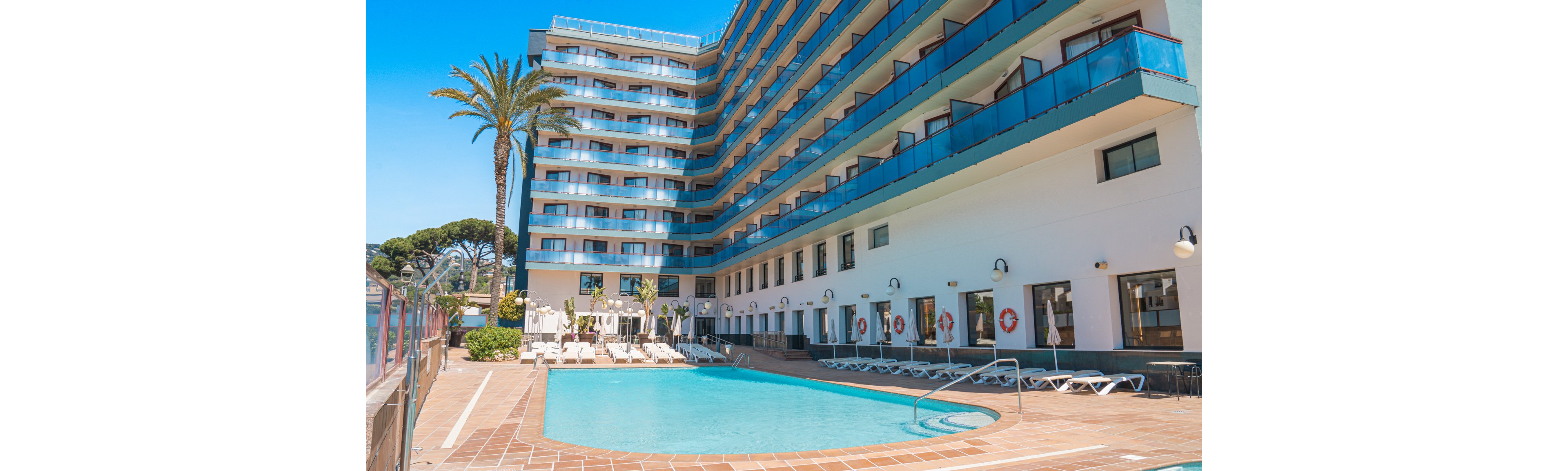 A multi-story hotel building features balconies overlooking a spacious outdoor swimming pool. The pool area is surrounded by sun loungers and palm trees, creating a relaxing vacation atmosphere. The scene is brightly lit with clear blue skies, and the pool water appears inviting. No visible text or numbers are present in the image.