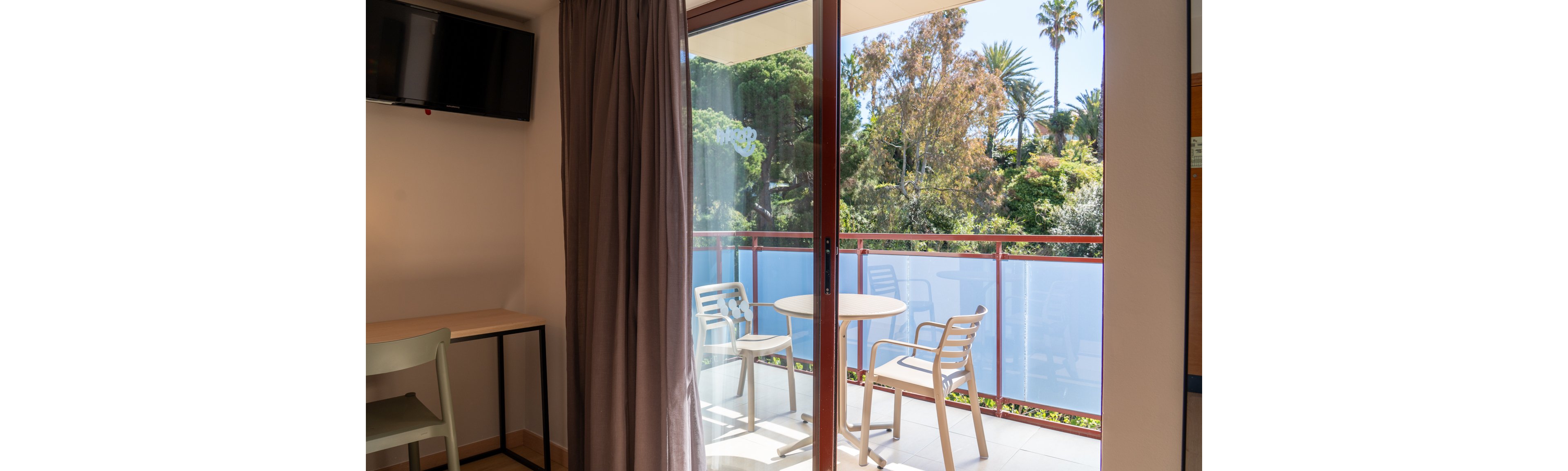 A bright hotel room features a small desk, chair, and wall-mounted television. Through the sliding glass door, a balcony with a round table and two chairs overlooks lush greenery and palm trees. Sunlight streams in, creating a relaxed and inviting atmosphere with neutral tones and natural light.