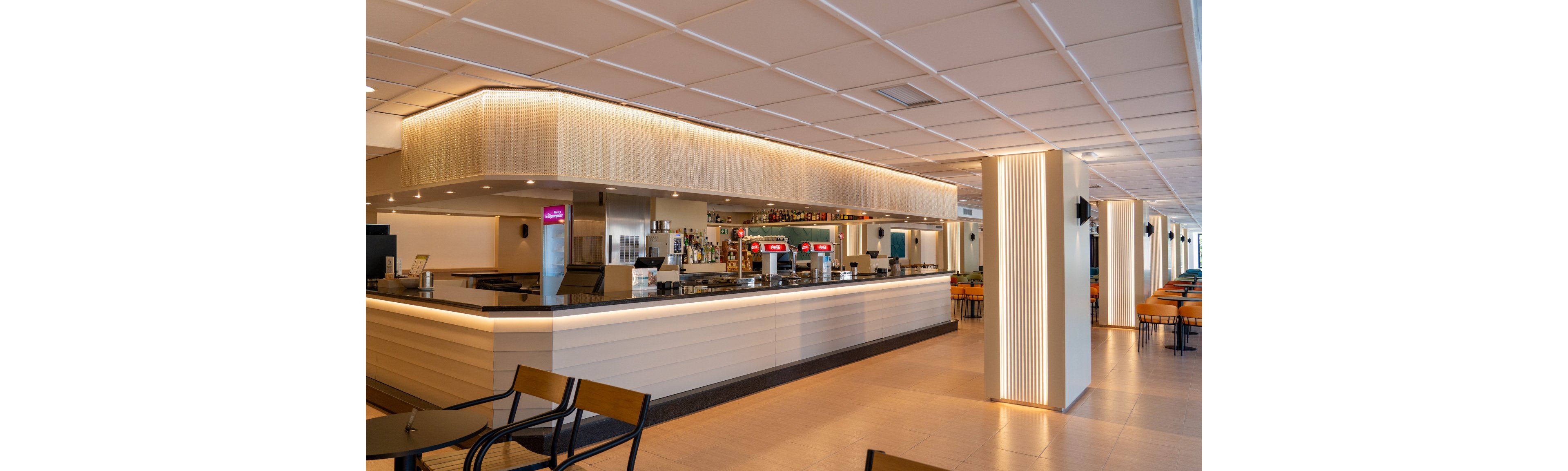 The image shows a contemporary bar area with a sleek, illuminated counter and several beverage dispensers. The setting is spacious and well-lit, featuring clean lines and neutral tones. Rows of tables and chairs are arranged neatly, and the overall mood is calm and inviting. No visible people or explicit numeric values are present in the scene.