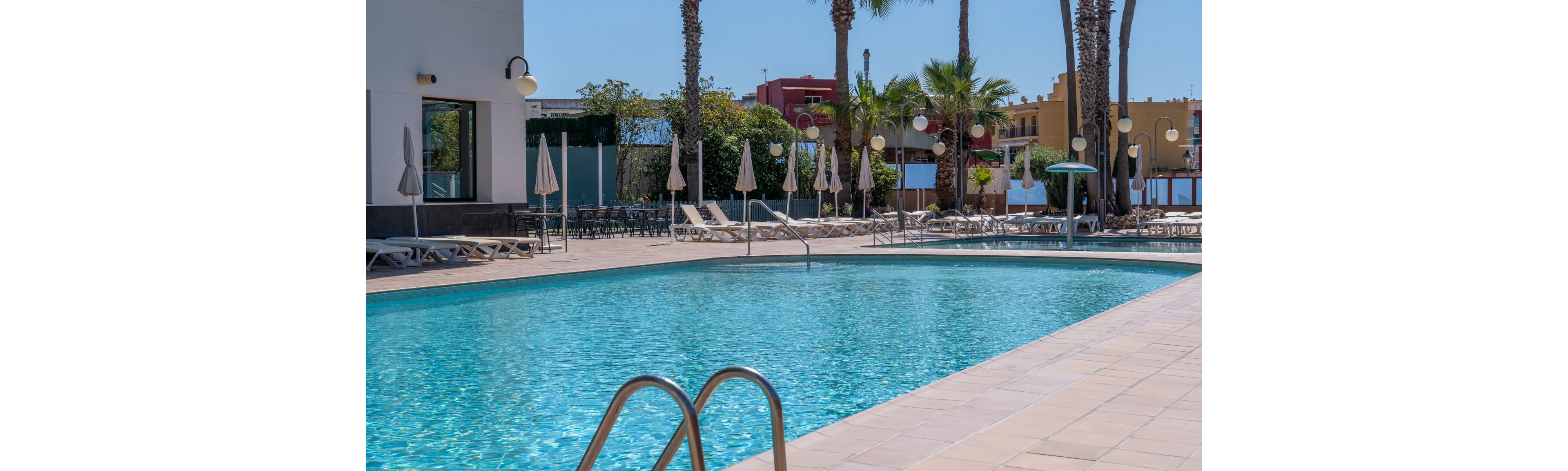 The main subject is a clear blue outdoor swimming pool surrounded by sun loungers and beige umbrellas. The setting is a sunny, resort-like area with palm trees and colorful buildings in the background. The mood is relaxed and inviting, with a bright, warm palette and clean visuals. No people are visible, emphasizing a tranquil atmosphere. No visible text or numeric values appear in the image.