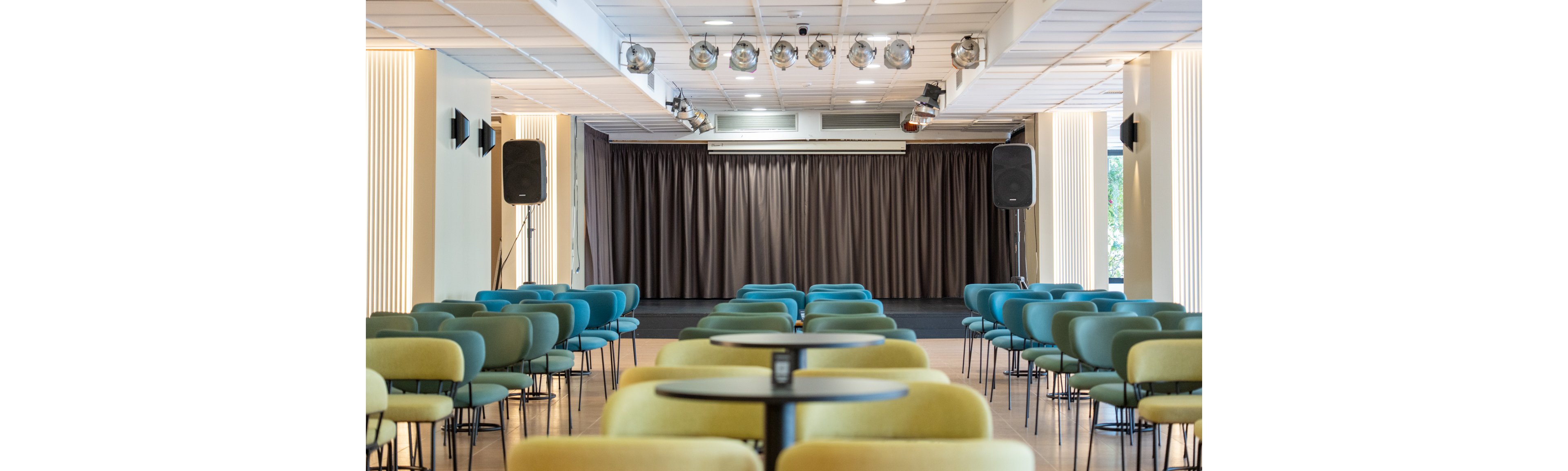 The image shows a contemporary auditorium featuring a stage with closed dark curtains and overhead lighting fixtures. Rows of blue and green chairs are arranged facing the stage, with round tables interspersed among the seating. Large speakers are positioned on either side of the stage, and the space is well-lit with vertical wall lights. The setting appears ready for a presentation, performance, or event.