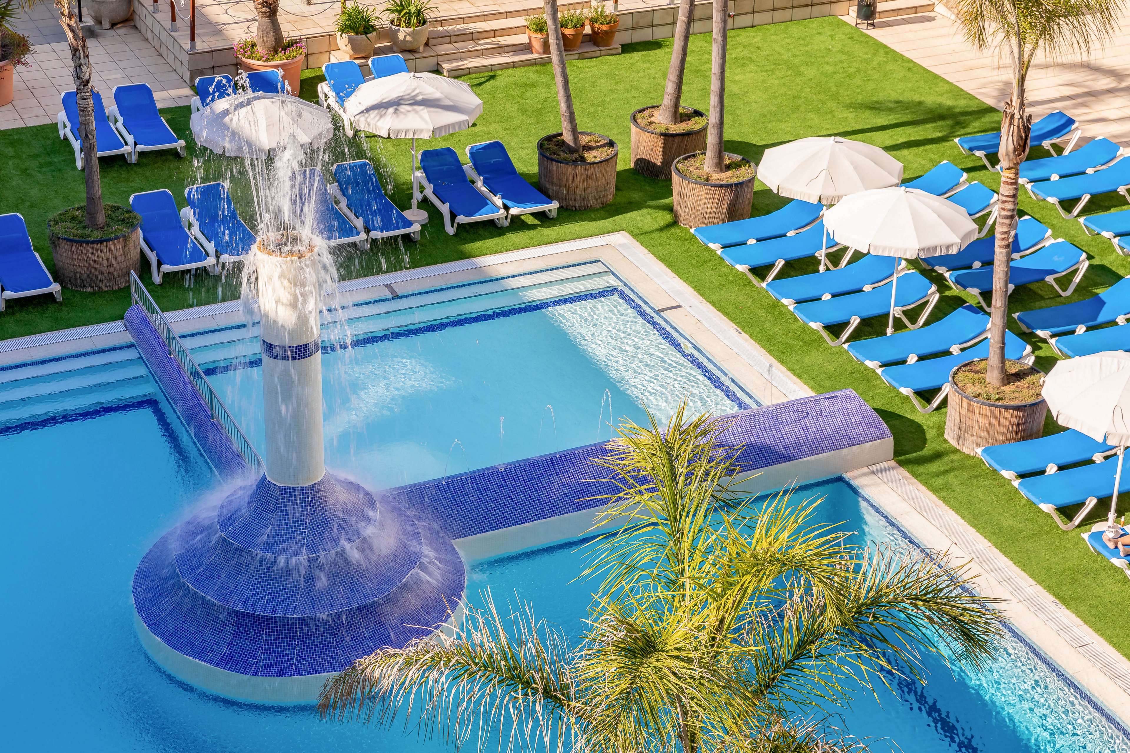 An outdoor resort swimming pool features a central fountain cascading water into the clear blue pool. Surrounding the pool are rows of blue lounge chairs and white umbrellas neatly arranged on a sun deck. Palm trees and green artificial grass add a tropical atmosphere to the leisure area. The bright sunlight and clean design create a relaxing vacation setting.
