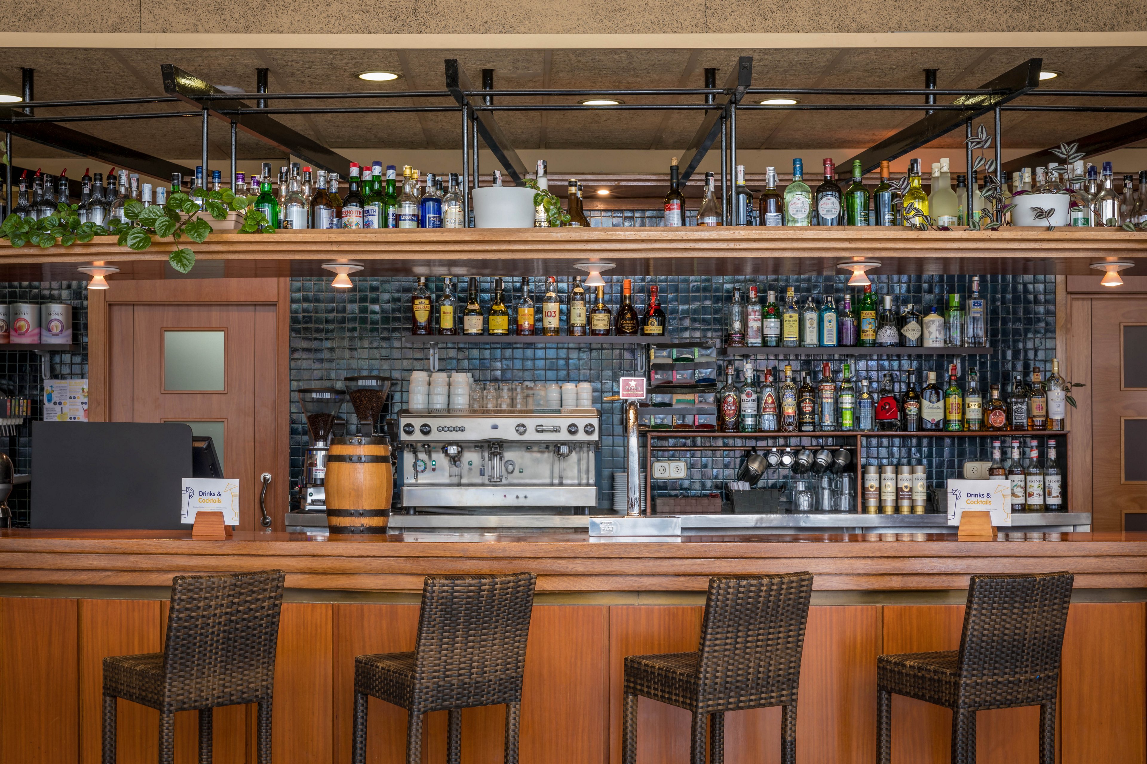 A bartender stands behind a polished wooden bar counter in a well-stocked lounge. Shelves filled with assorted liquor bottles and glassware line the wall behind him. He wears a white shirt, black vest, and tie while preparing a drink at the center of the bar. Four wicker bar stools are arranged neatly in front of the counter, and small table signs sit on the bar surface.