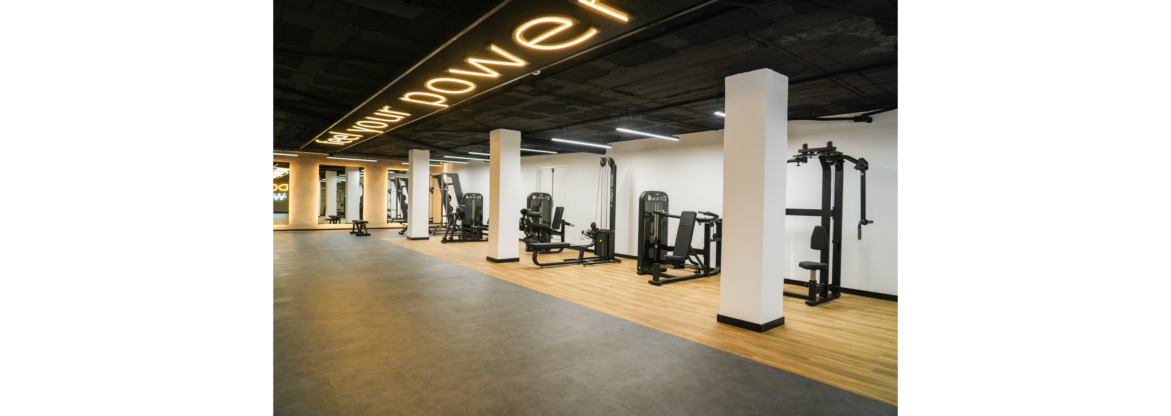 A spacious, modern gym interior features several strength training machines arranged in neat rows. The room has a combination of wood-style flooring and darker rubber flooring, creating distinct workout zones. Bright white columns and walls contrast with the dark ceiling, where illuminated text signage adds a stylish accent. The gym is empty, emphasizing clean lines, open space, and contemporary fitness design.
