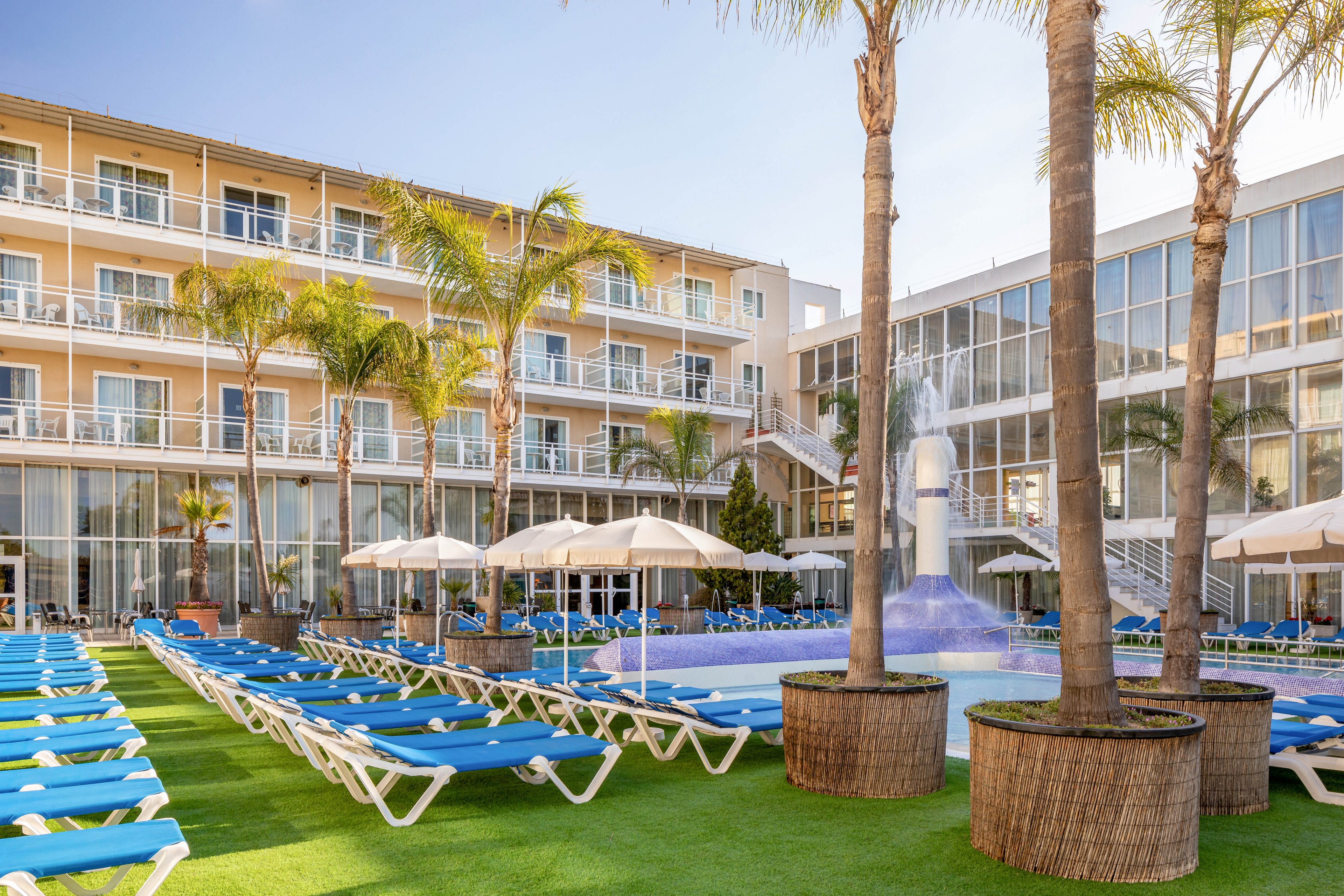 An outdoor hotel courtyard features a swimming pool surrounded by rows of blue sun loungers. Tall palm trees and large potted plants add a tropical feel to the modern multi-story buildings. White umbrellas provide shaded seating areas around the pool. The bright daylight and clear sky create a warm, inviting vacation atmosphere.