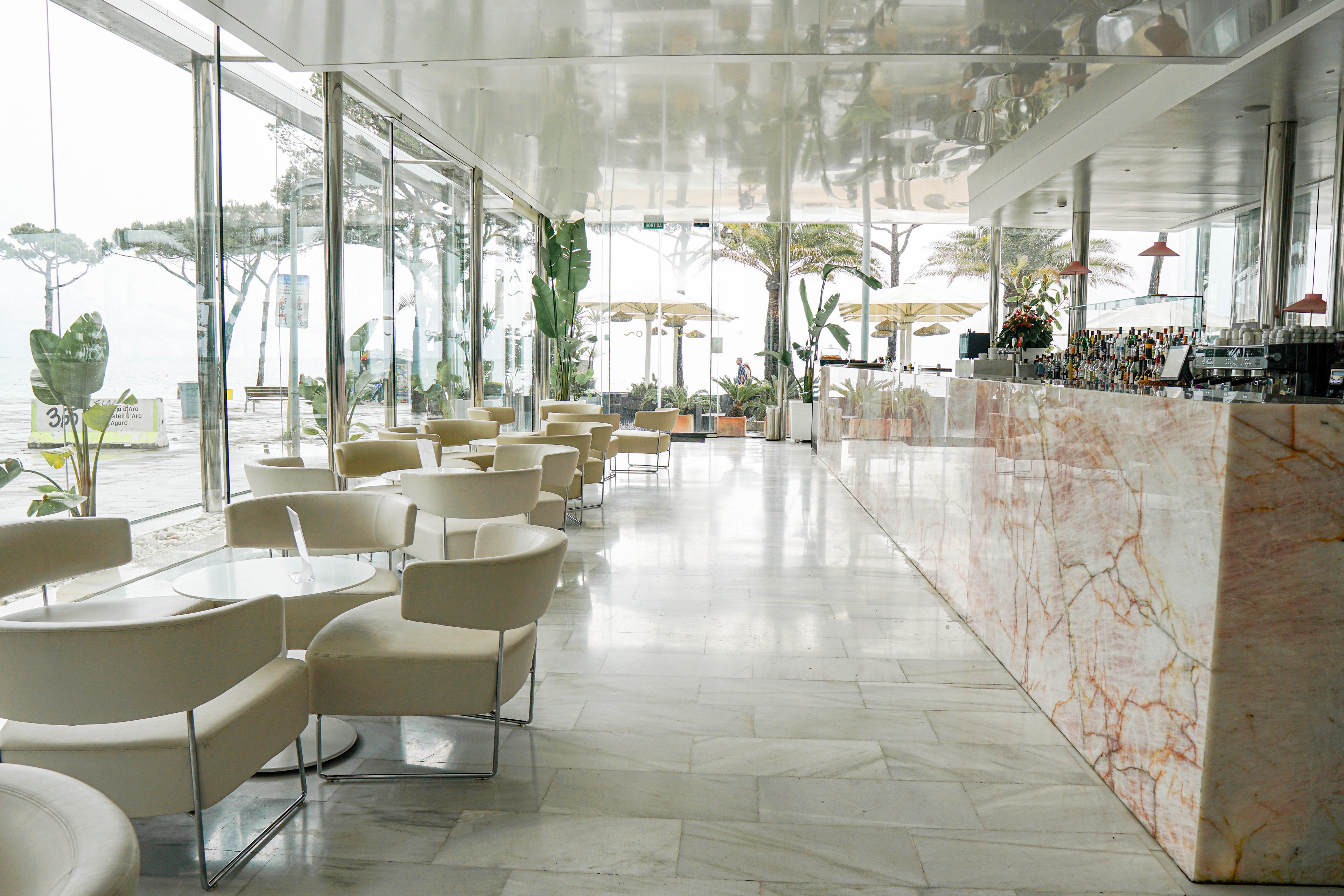 A contemporary café interior features cream-colored armchairs and round tables arranged neatly along a glass wall. Large floor-to-ceiling windows reveal a beachfront view with trees and outdoor umbrellas. A long marble bar counter runs along the right side, displaying bottles and bar equipment. The space is bright, airy, and minimalist, with reflective surfaces and a neutral color palette.
