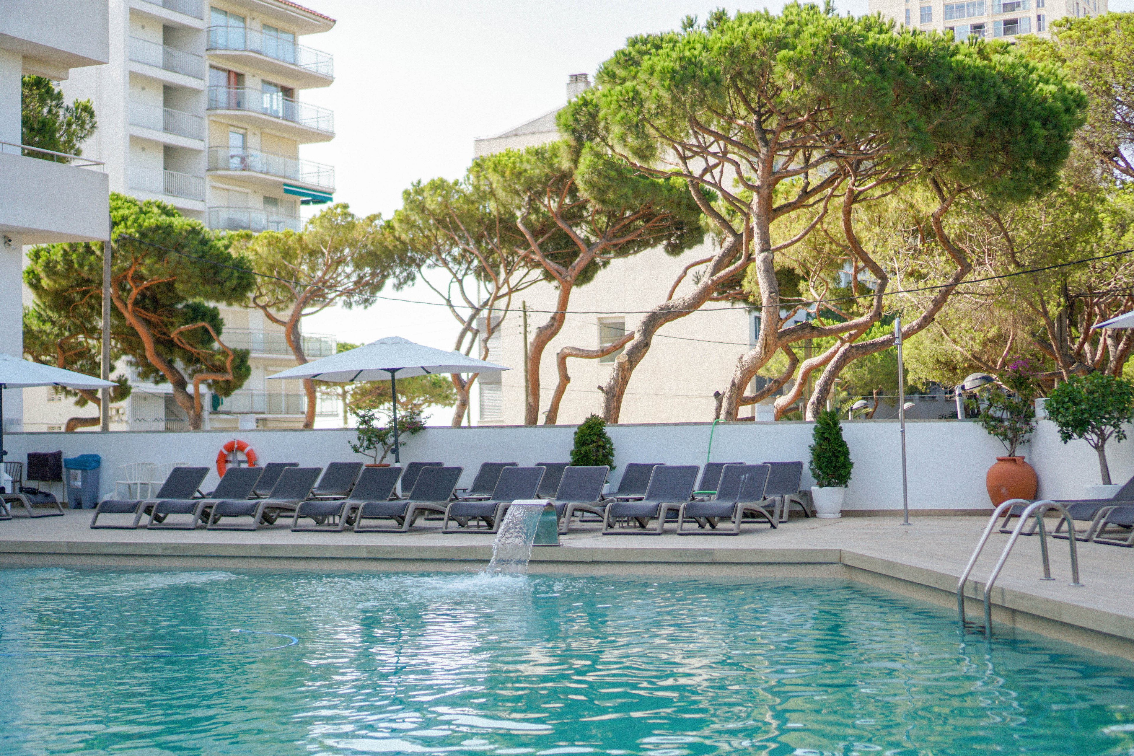 A modern outdoor swimming pool is surrounded by rows of empty sun loungers and umbrellas. Clear turquoise water with a small fountain feature dominates the foreground. Tall sculpted trees provide natural shade behind a white wall, with potted plants adding greenery. A multi-story residential or hotel building rises in the background, suggesting an urban resort setting. The scene feels calm and inviting, ideal for relaxation and leisure.