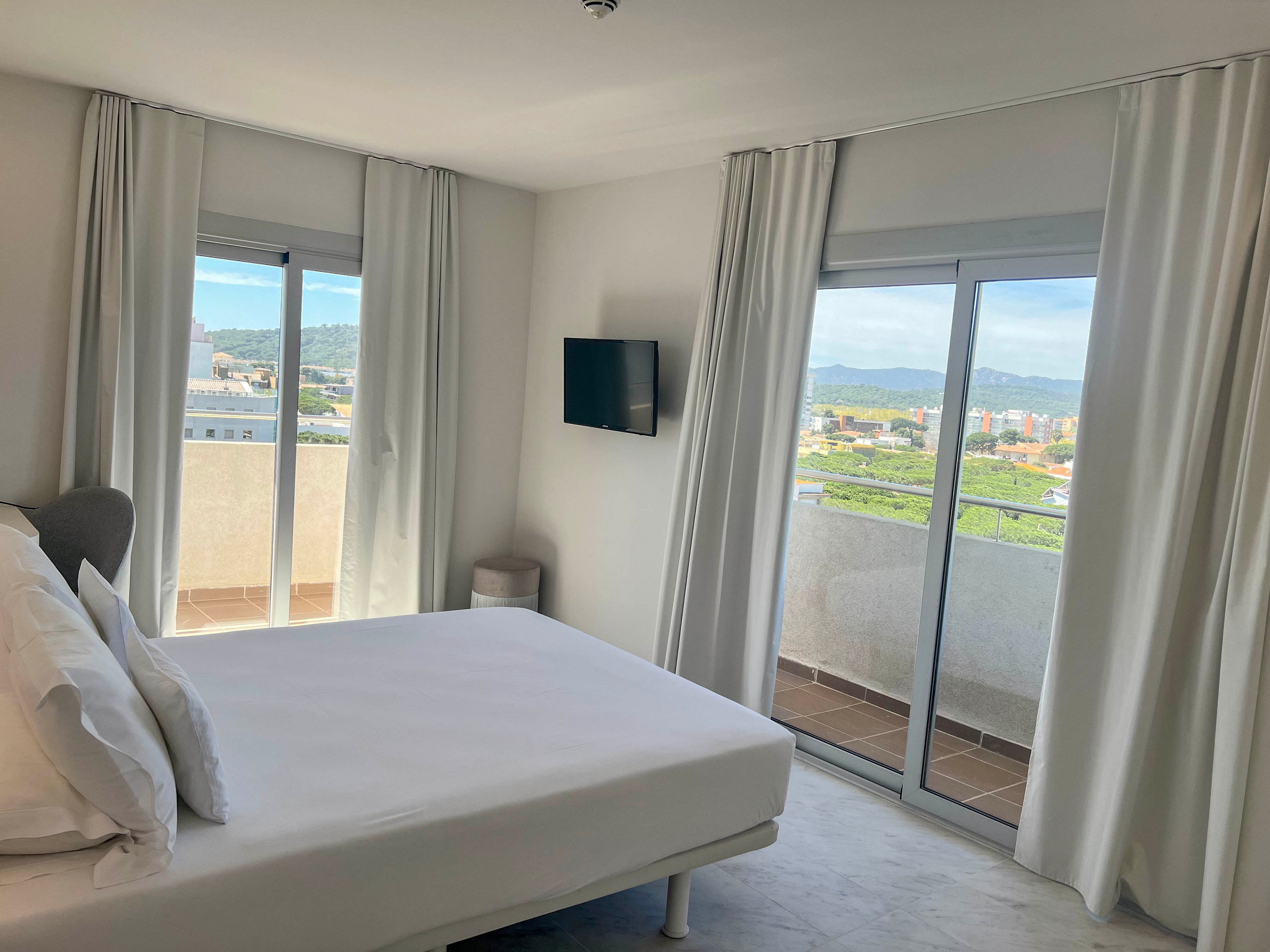 A modern hotel bedroom features a neatly made bed with white linens and pillows. Large sliding glass doors open onto balconies, offering a scenic view of a city and distant mountains. White curtains frame the windows, enhancing the bright and airy atmosphere. A flat-screen television is mounted on the wall, adding a contemporary touch to the minimalist decor.