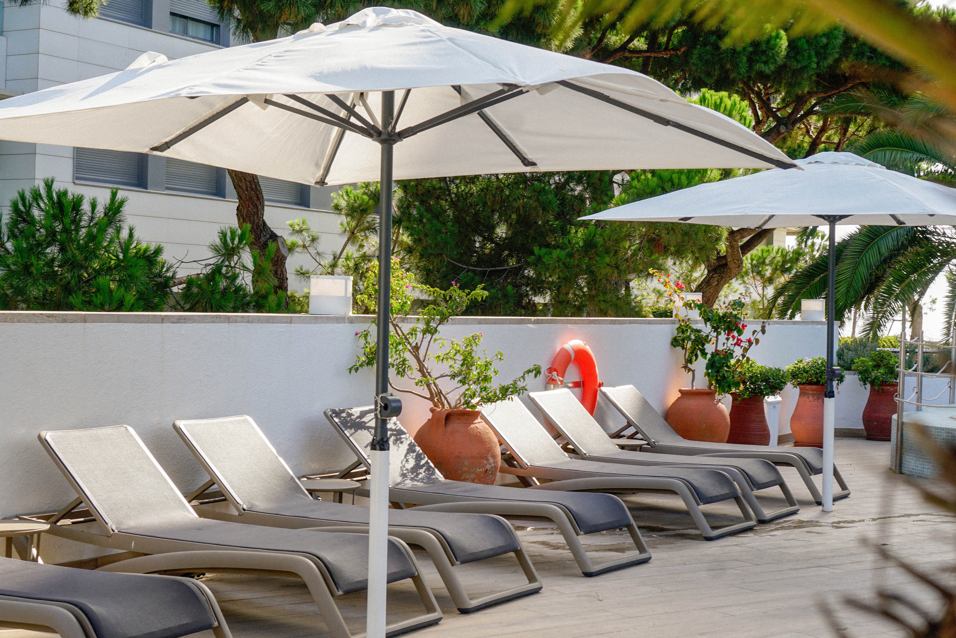 A row of modern gray lounge chairs is arranged neatly on a sunlit terrace. Large white umbrellas provide shade along the deck, creating a relaxing poolside atmosphere. Terracotta pots with green plants and a bright orange lifebuoy add color against the white wall. Trees and a building facade in the background suggest a resort or hotel setting.