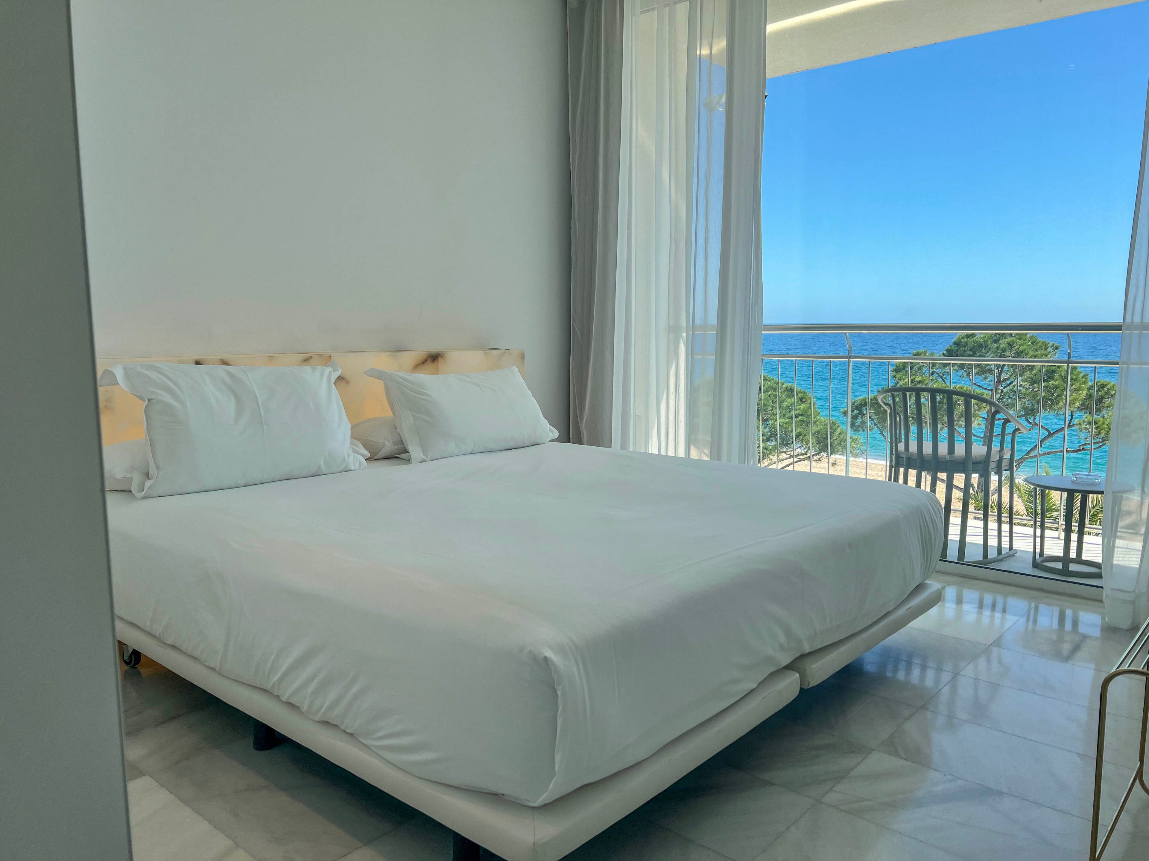 A modern hotel bedroom features a large neatly made bed with crisp white linens. Floor-to-ceiling windows and sheer curtains open onto a balcony overlooking a bright blue sea and green coastal trees. Natural light fills the minimalist room, highlighting the clean white walls and glossy floor. A simple chair and table on the balcony invite guests to relax and enjoy the ocean view.