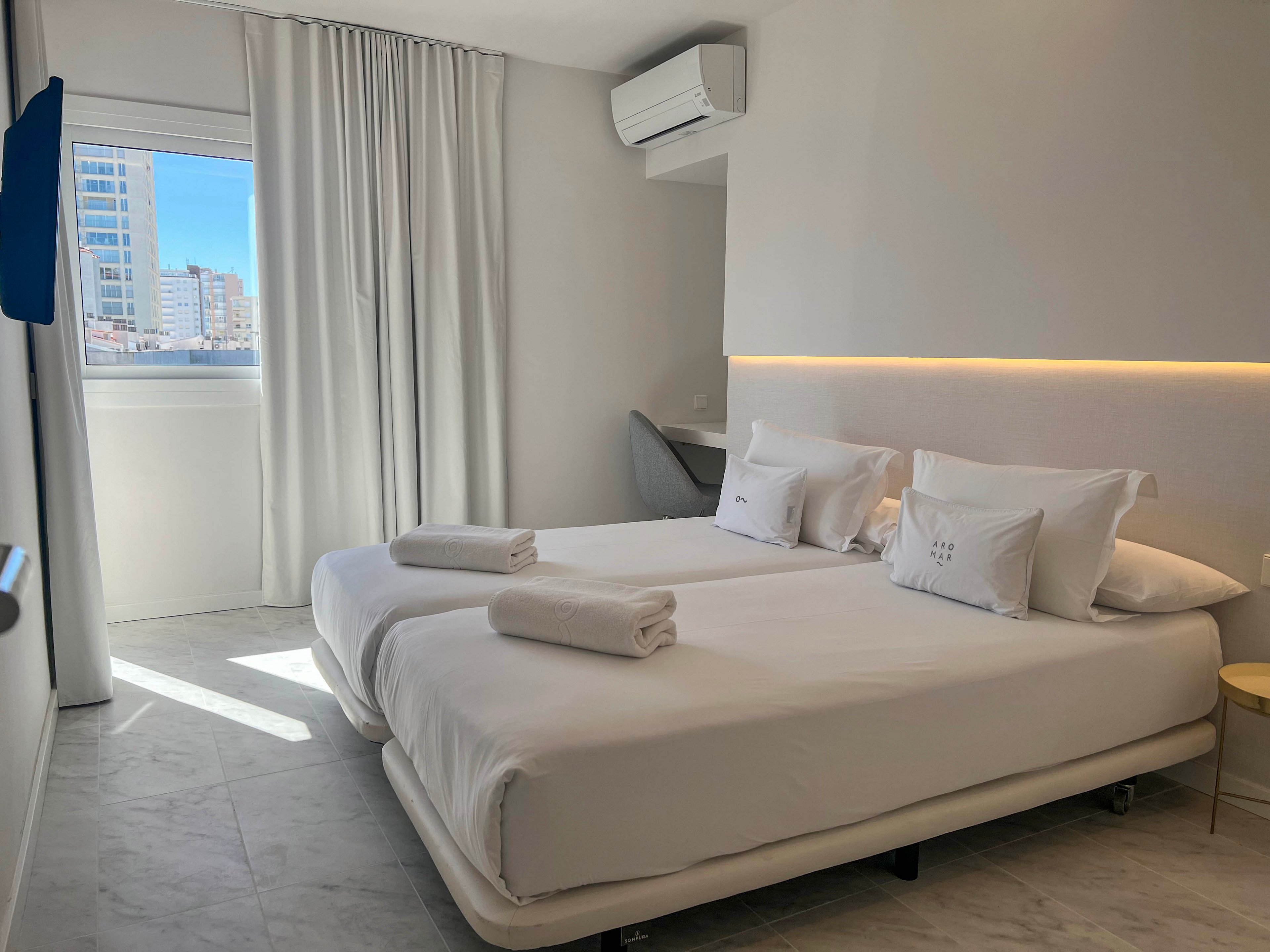 A bright, modern hotel room features two neatly made twin beds pushed together, each topped with folded white towels and pillows. Natural light streams in through a large window with sheer white curtains, revealing a cityscape of tall buildings outside. The room has a minimalist design with white walls, a marble-look floor, and a subtle strip of warm accent lighting above the headboard. An air conditioning unit and a small desk with a chair are visible, emphasizing comfort and functionality in the compact space.