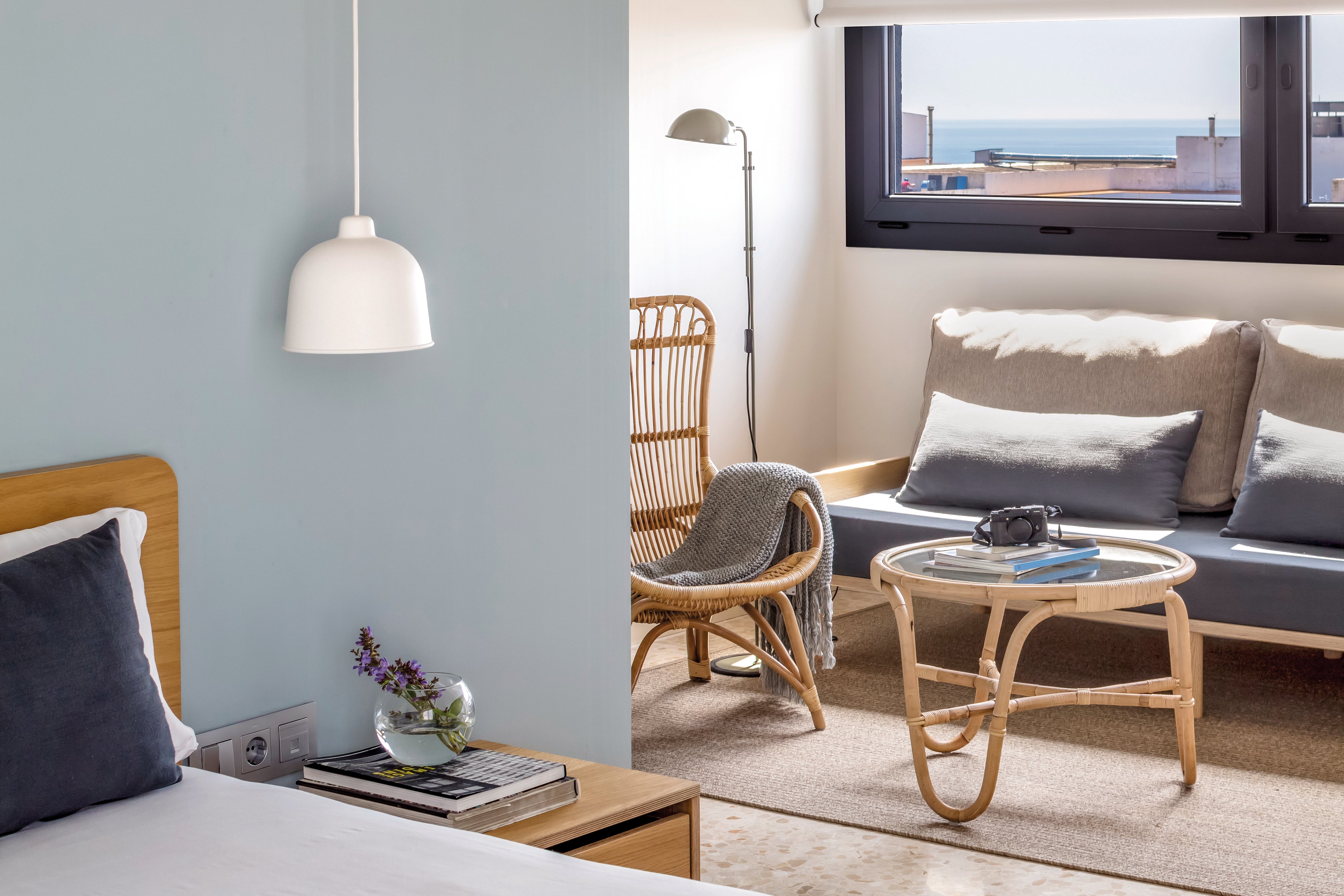 A bright and cozy room featuring a wooden bed and a seating area with a rattan chair and sofa. The setting includes soft neutral tones and a view of the ocean through large windows. Decorative elements include a pendant lamp, a small vase with flowers, and a camera on a glass-top table.