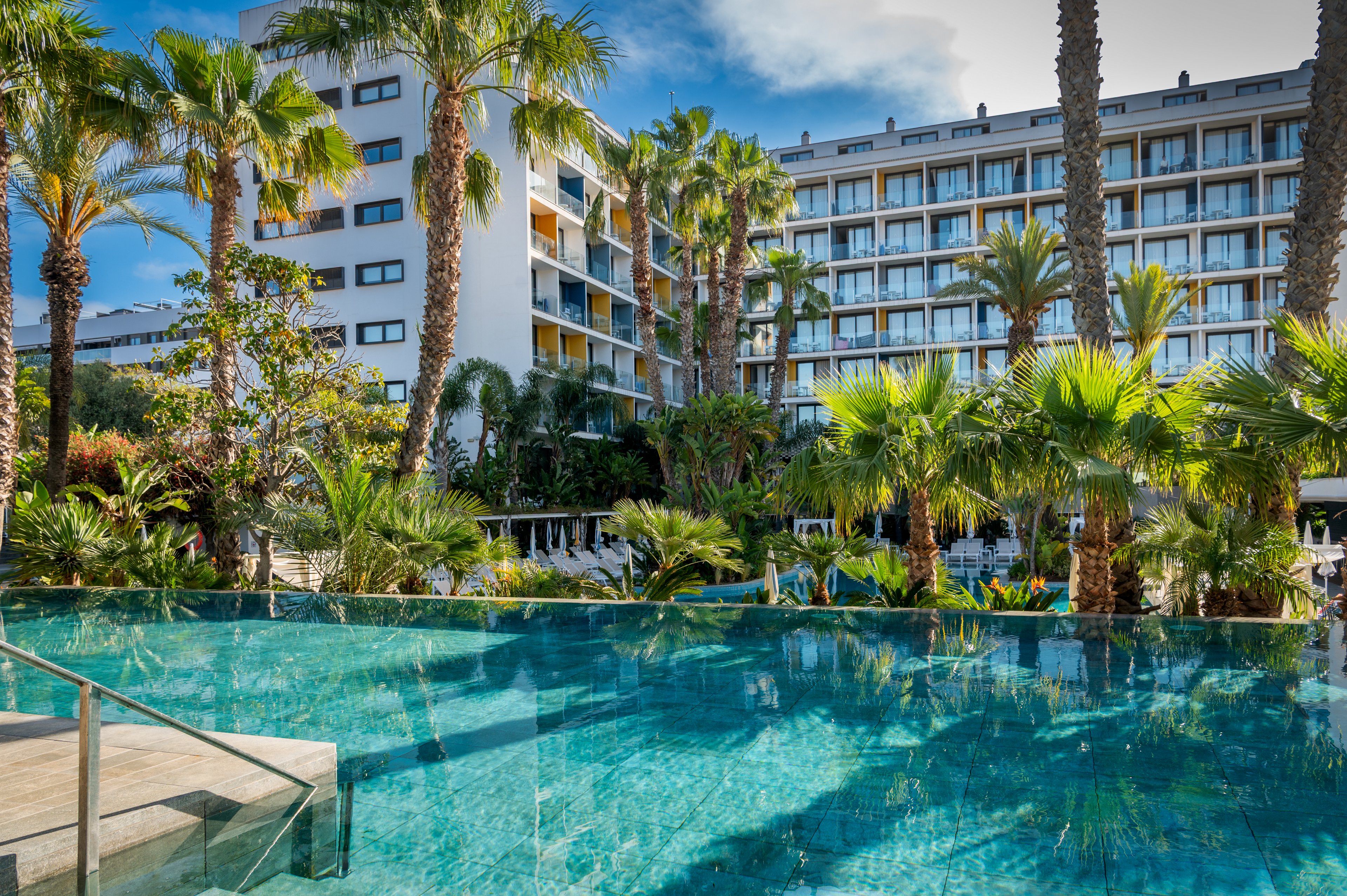 A modern multi-story resort hotel overlooks a clear blue infinity pool. Tall palm trees and lush tropical plants surround the water, creating a relaxing vacation atmosphere. Sun loungers and umbrellas are visible in the background near the pool area. The bright daylight and blue sky enhance the fresh, inviting feel of the outdoor space.