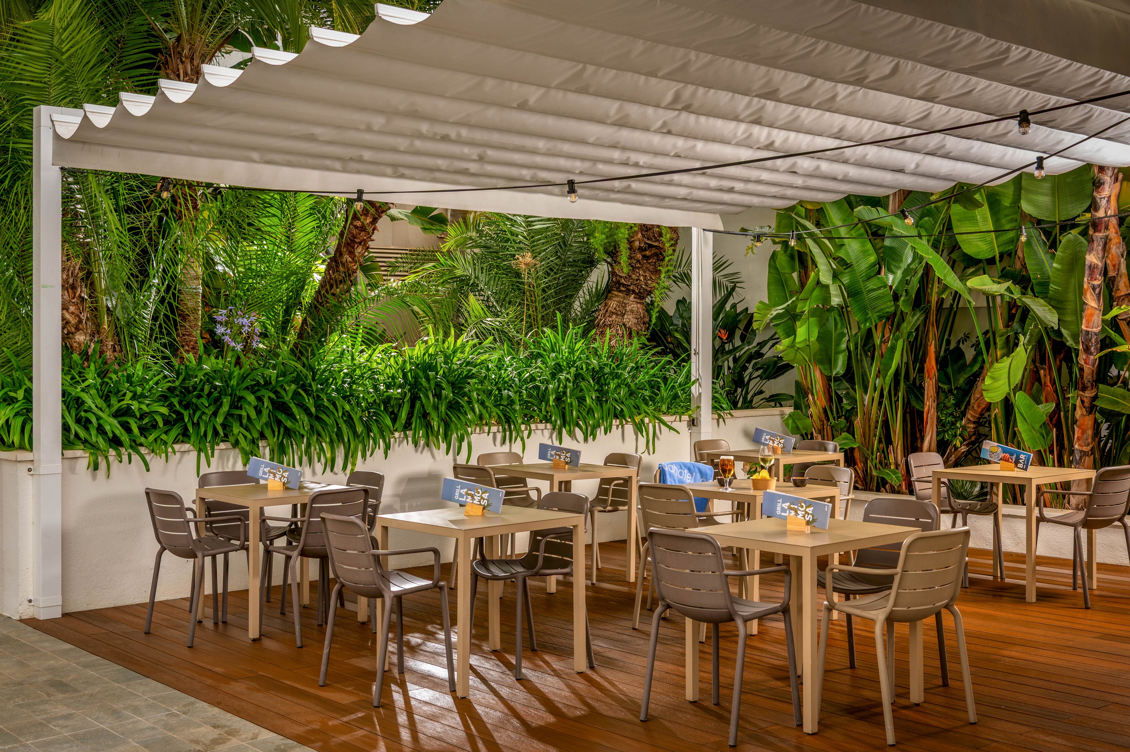 An outdoor dining terrace features several empty tables and chairs arranged neatly under a white pergola-style canopy. The setting is surrounded by lush tropical plants and palm trees, creating a private and relaxing atmosphere. Warm lighting on the tables suggests a welcoming space for evening meals or casual gatherings. The wooden deck flooring contrasts with the greenery and enhances the cozy, resort-like ambiance.