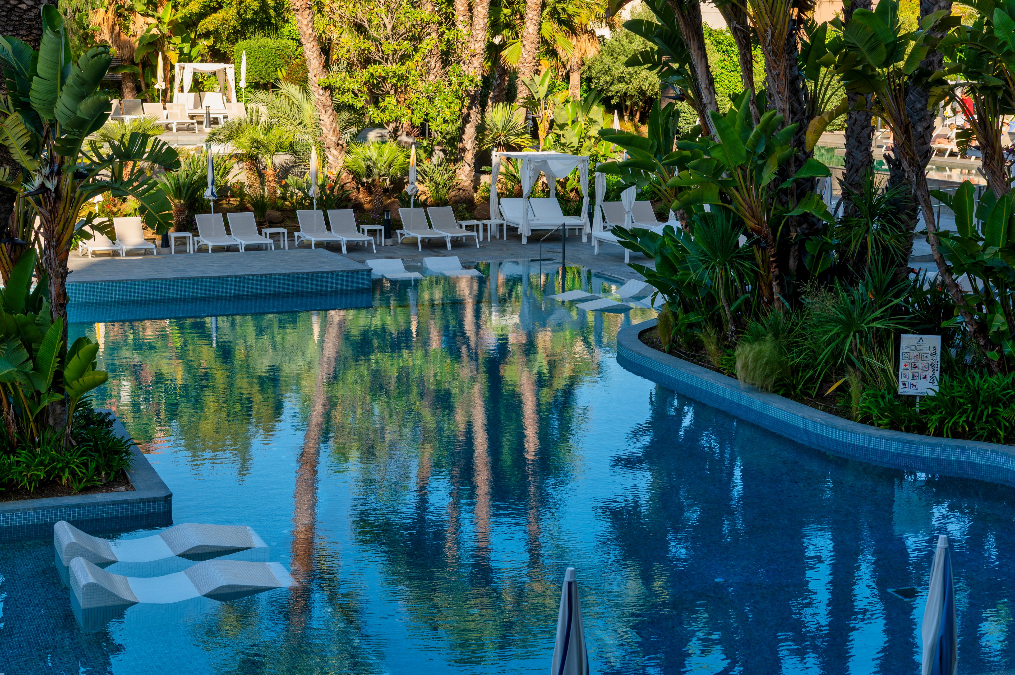 A tranquil resort swimming pool is surrounded by lush tropical greenery and tall palm trees. White sun loungers and cabanas line the poolside, suggesting a relaxing vacation setting. Two modern white pool loungers float on the calm blue water, reflecting the surrounding plants and sky. The overall scene feels serene and luxurious, ideal for leisure and sunbathing.