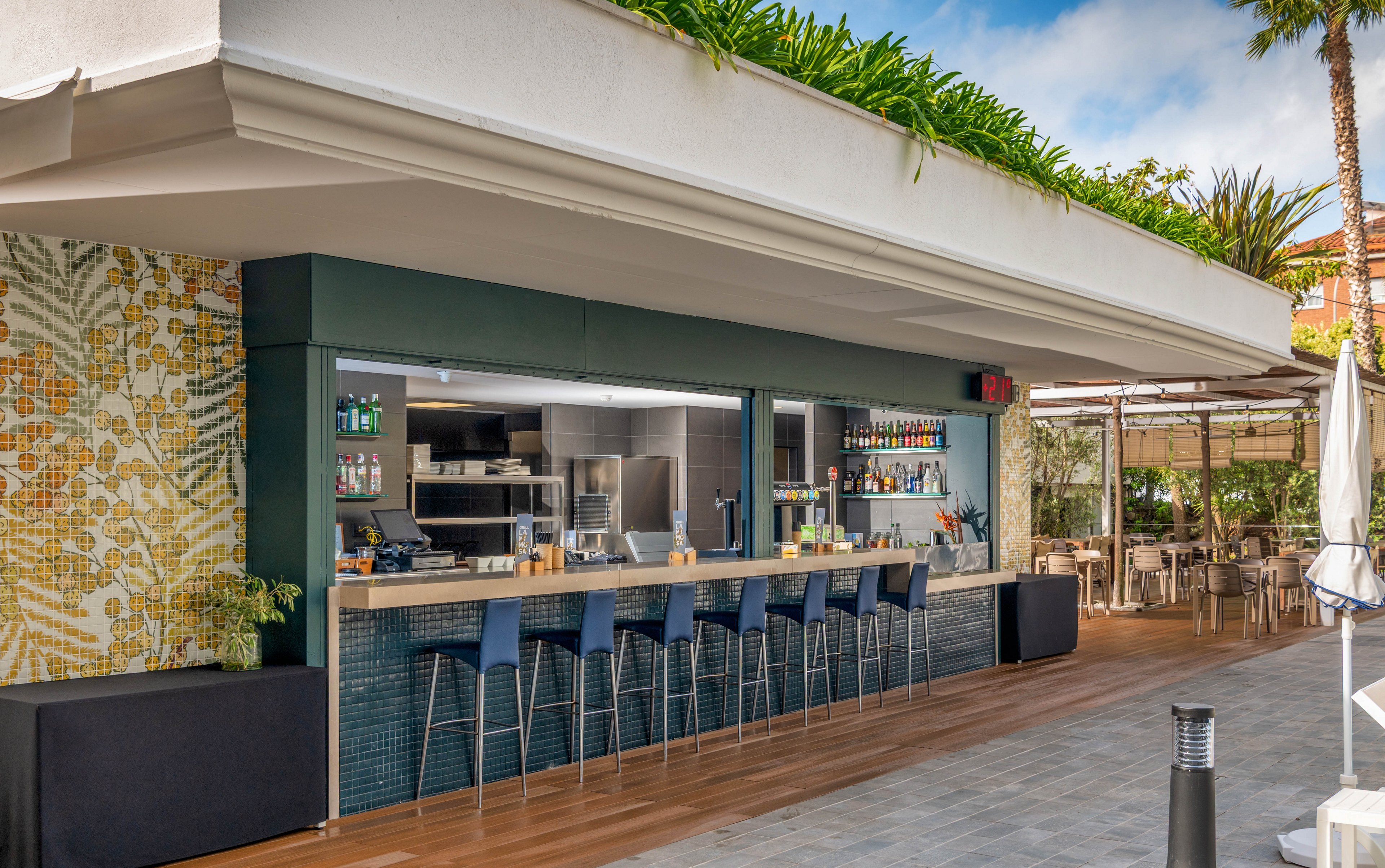 Open-air bar with a long counter and high stools is set beneath a modern overhang. The bar shelves display bottles and glassware, with a compact kitchen visible in the background. A mosaic wall, wooden deck flooring, and surrounding greenery create a relaxed resort atmosphere. The scene is bright and inviting, suggesting a casual vacation or poolside dining experience.
