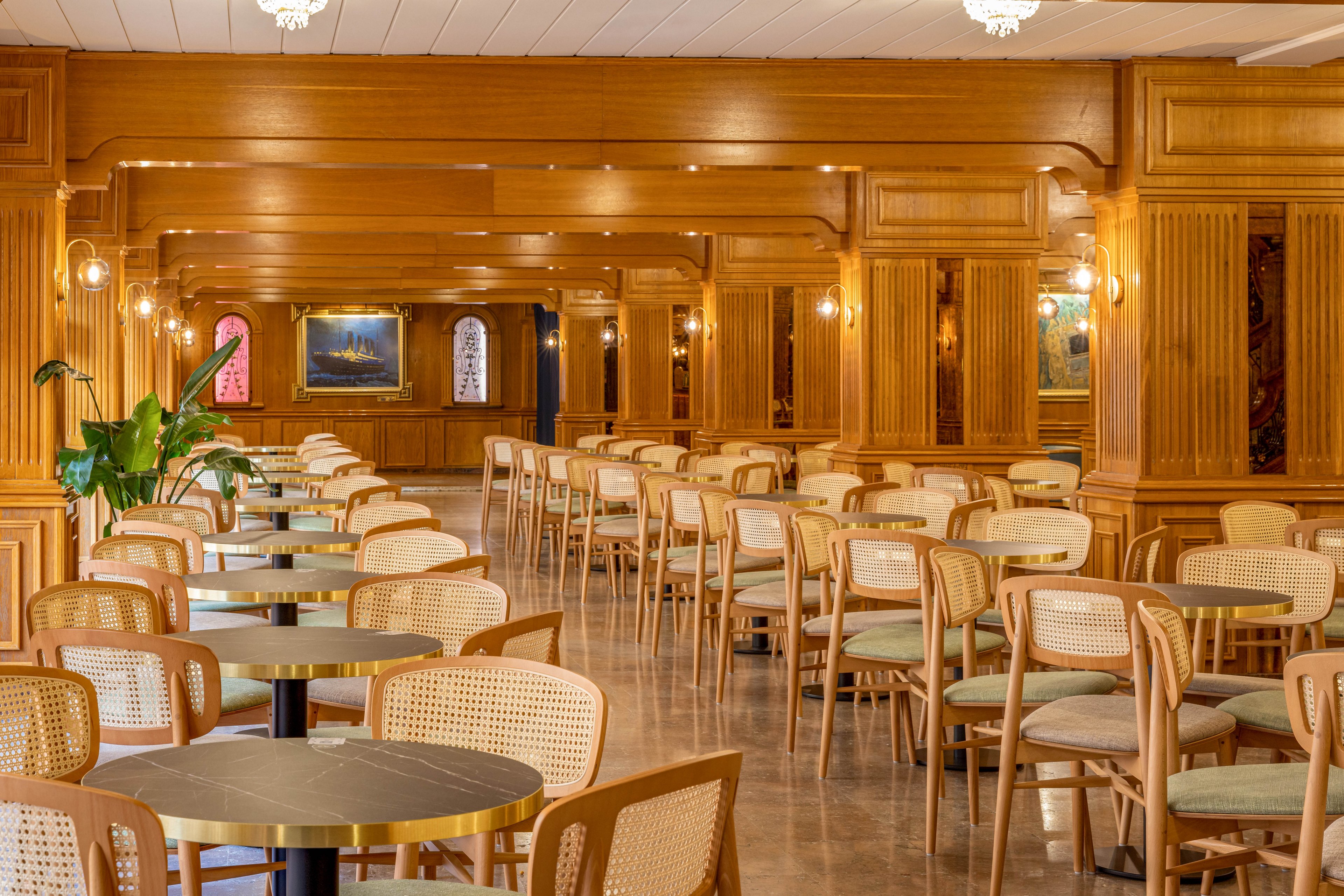 Spacious restaurant interior with rows of light wooden tables and woven-back chairs arranged neatly. Warm wood paneling covers the walls and columns, complemented by soft wall sconces and ceiling lights. Framed artwork and arched wall openings add visual interest to the background. A potted green plant introduces a touch of nature to the polished, reflective floor. The room appears ready for guests, creating a calm and inviting atmosphere.