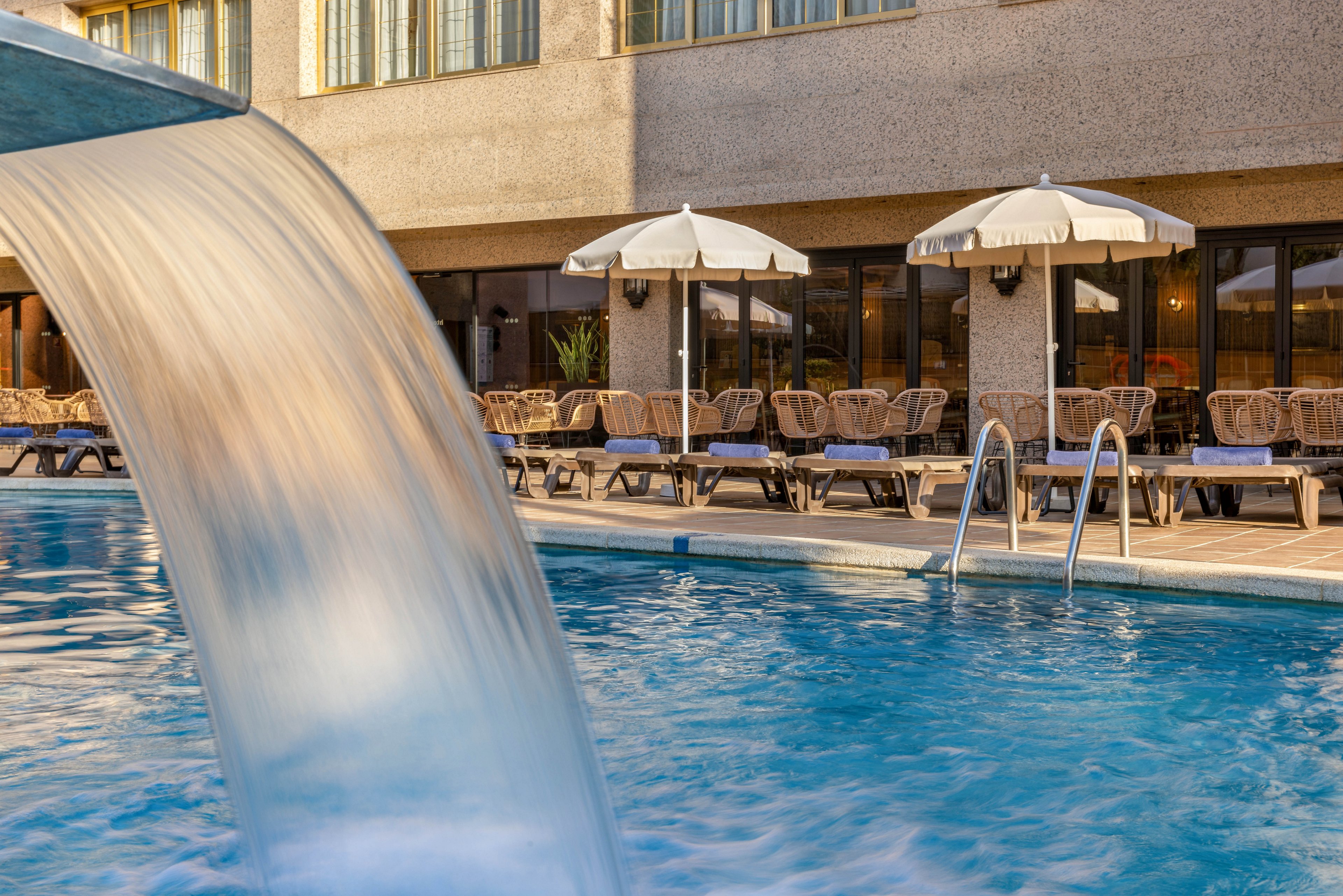 An outdoor swimming pool features a curved water fountain in the foreground. In the background, rows of lounge chairs and white umbrellas line the poolside terrace. The setting appears to be part of a hotel or resort building with large windows and a textured facade. The scene suggests a calm, sunny day ideal for relaxation by the water.