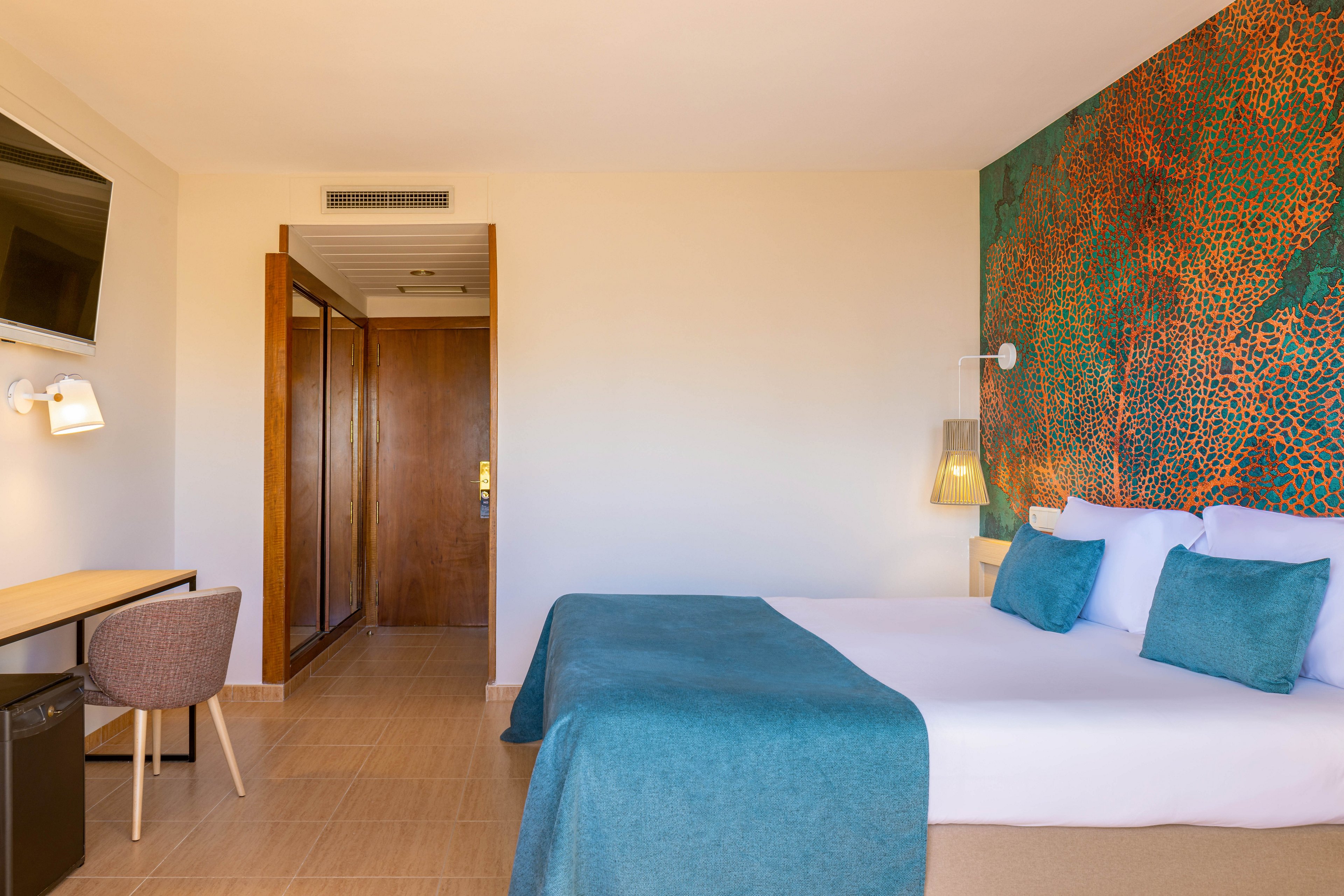 A neatly made double bed with teal bedding and pillows is the focal point of this modern hotel room. The space features a colorful abstract wall mural behind the bed and warm wooden doors leading to the hallway or closet area. A small desk with a chair, wall-mounted television, and bedside lighting complete the contemporary interior. The room appears bright, clean, and inviting, with neutral walls and tiled flooring.