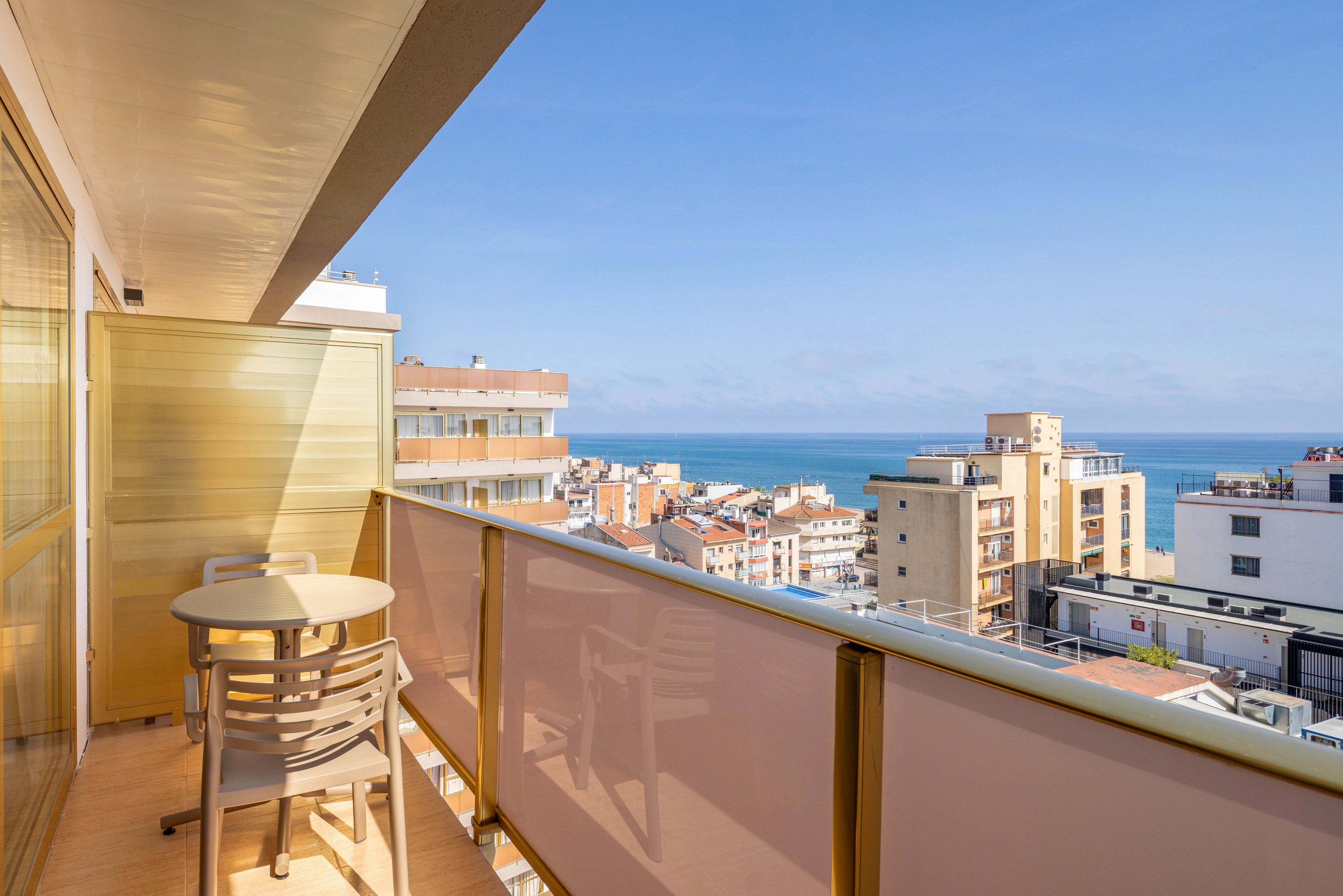 A bright hotel balcony with a small round table and two chairs overlooks a coastal cityscape. Modern apartment and hotel buildings stretch toward the horizon under a clear blue sky. In the distance, the calm sea creates a serene backdrop to the urban setting. Warm sunlight enhances the relaxed, vacation-like atmosphere of the scene.