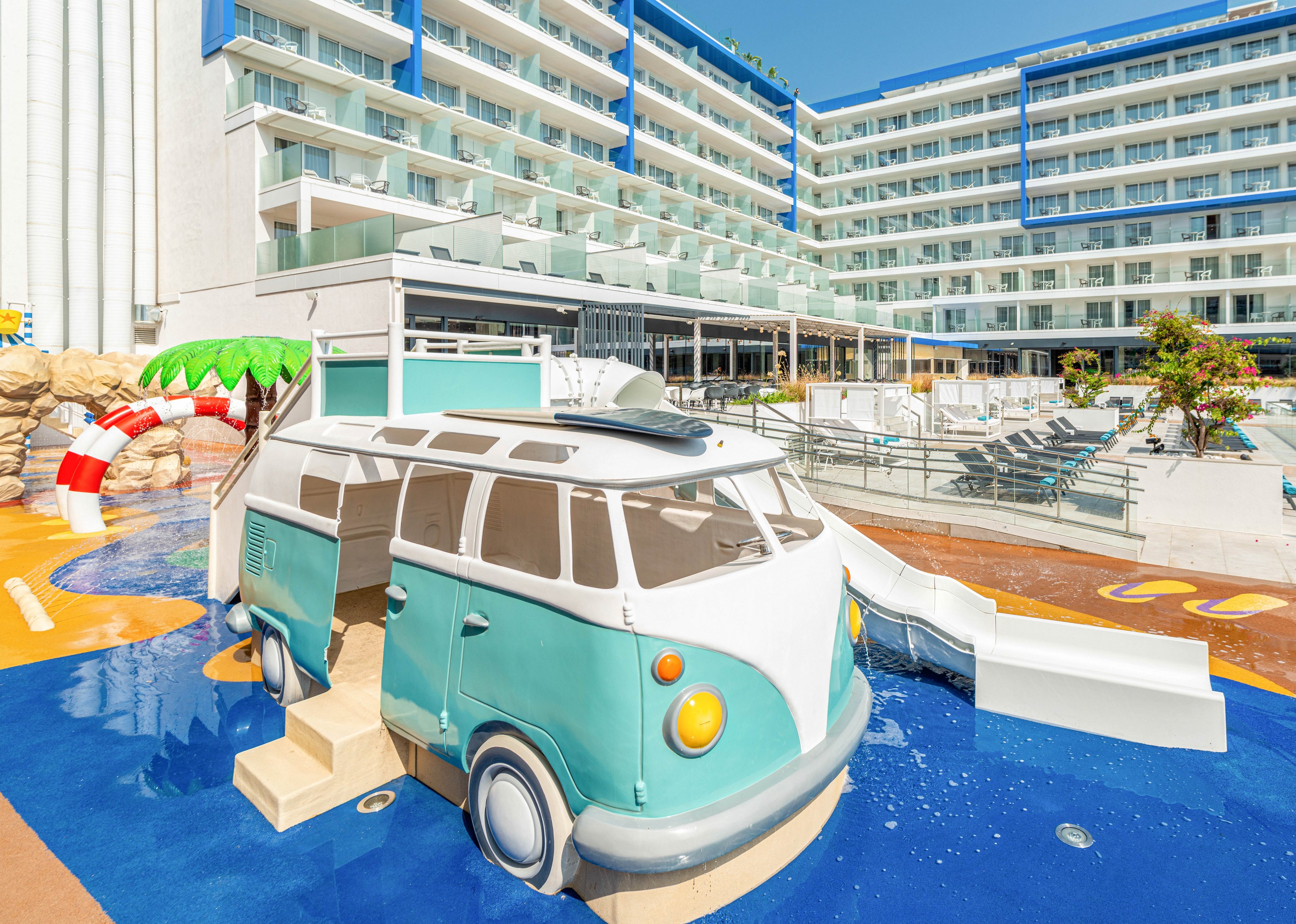 A playful camper van–shaped water slide sits in a shallow children's pool at a modern resort hotel. The turquoise and white slide structure features steps, windows, and a curved body leading to a smooth white chute. In the background, a large multi-story hotel with balconies overlooks sun loungers and additional pool areas. Bright colors, clear skies, and clean architectural lines create a cheerful vacation atmosphere.