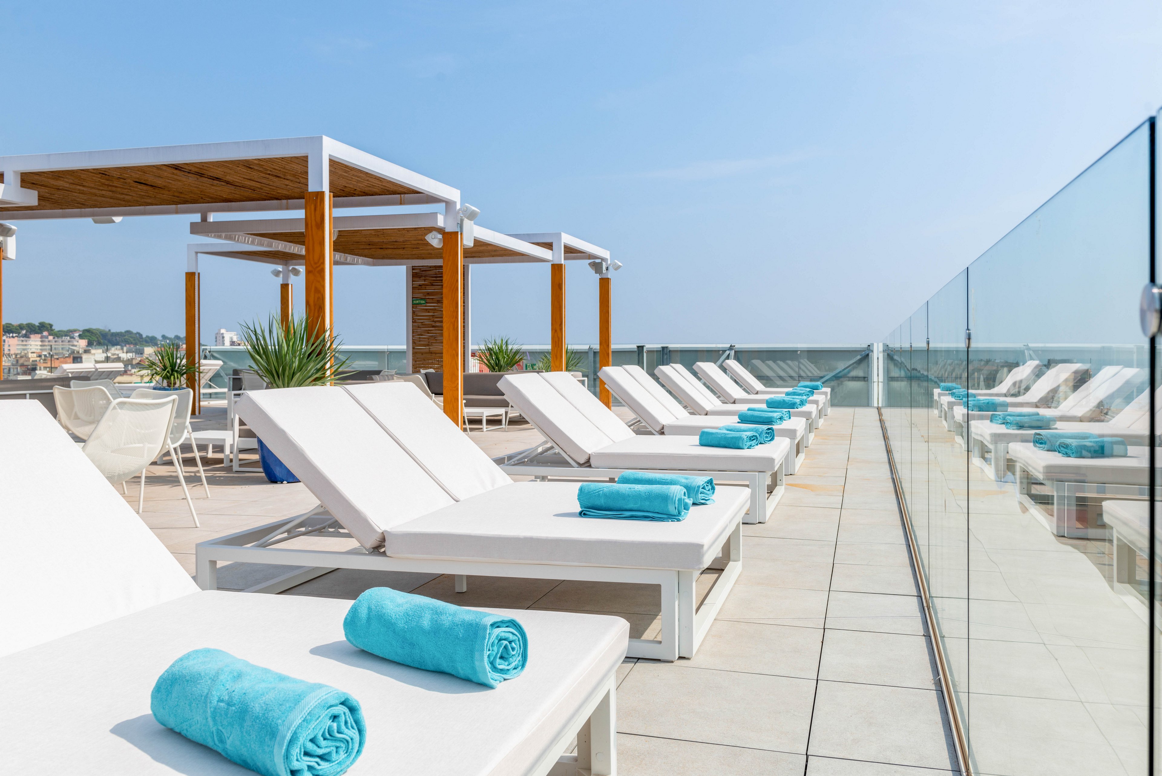 A row of white sun loungers with neatly rolled turquoise towels is arranged on a modern rooftop terrace. Glass railings line one side, offering an open view toward the distant cityscape under a clear blue sky. Wooden pergolas and potted plants add structure and greenery to the bright, minimalist setting. The scene suggests a tranquil hotel pool area or spa designed for relaxation and sunbathing.