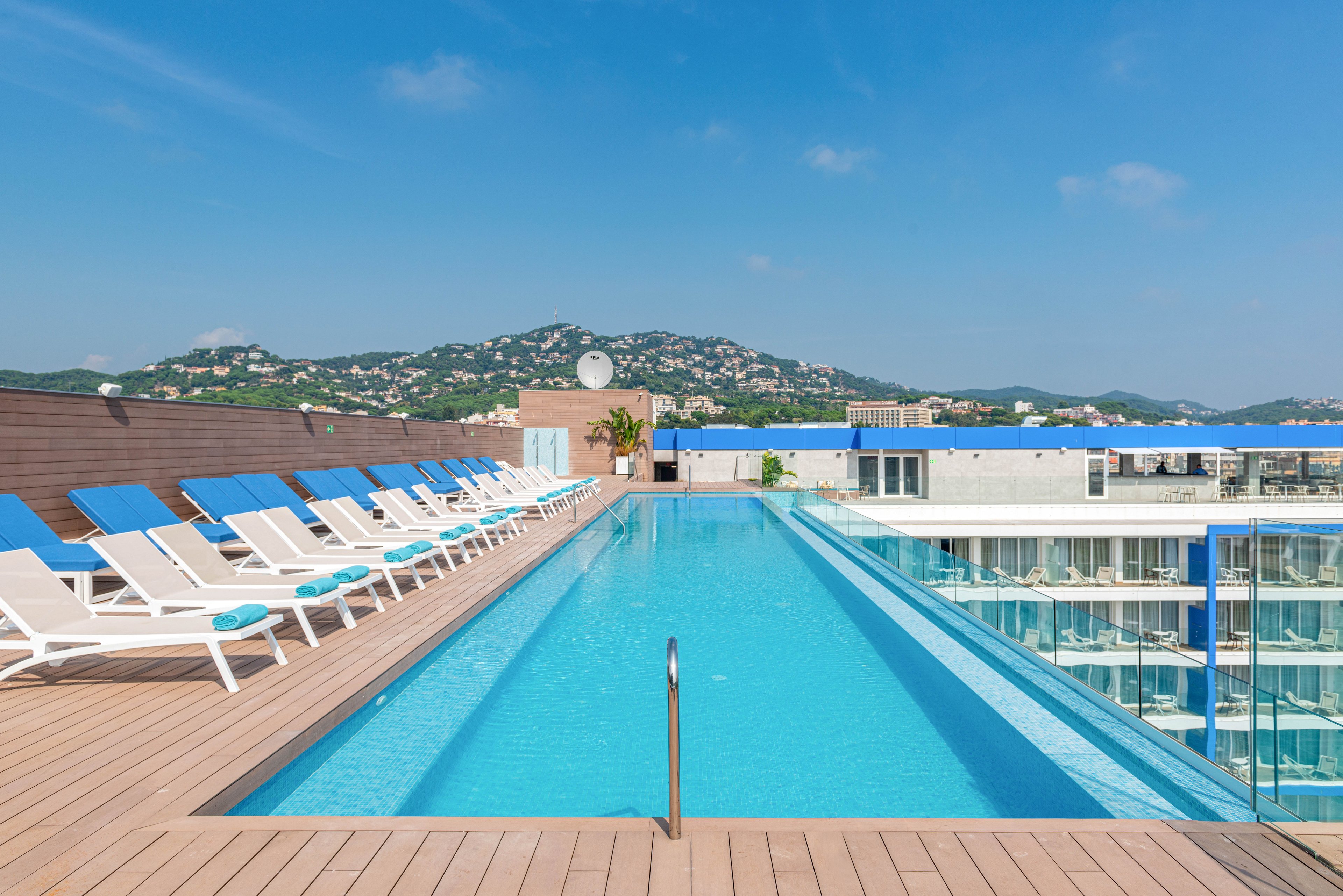 A long rooftop infinity pool stretches between rows of white loungers with blue cushions. The pool sits atop a modern hotel building with glass balconies and clean architectural lines. In the distance, green hills dotted with houses rise under a clear blue sky. The bright sunlight and calm water create a relaxing, resort-like atmosphere ideal for vacation or leisure.