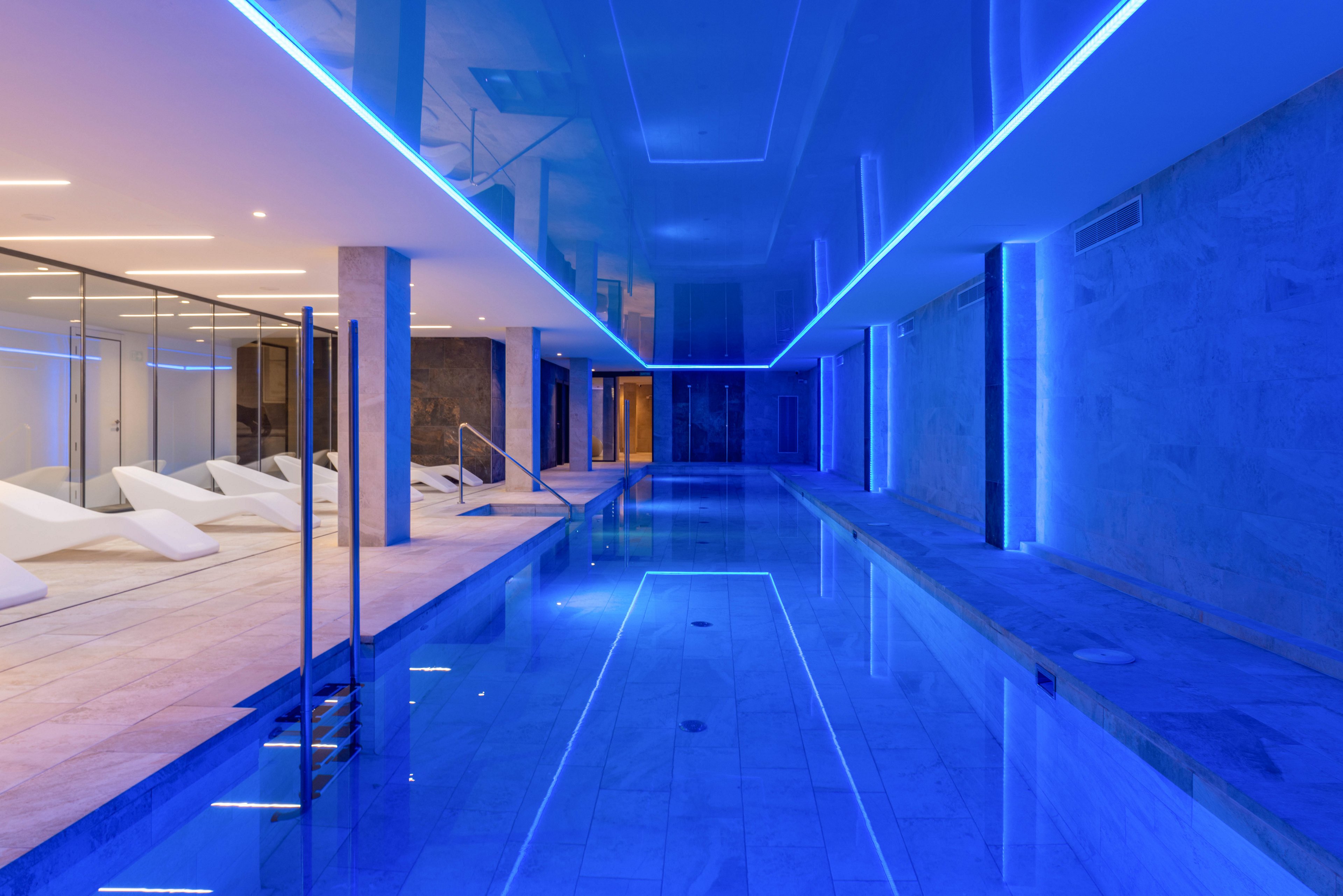 A sleek indoor swimming pool is illuminated by vibrant blue LED strip lighting along the ceiling and walls. The space features clean architectural lines, glass partitions, and stone surfaces for a contemporary spa-like atmosphere. White lounge chairs are arranged behind the glass, suggesting a relaxation area adjacent to the pool. The overall scene feels calm, luxurious, and minimalist.