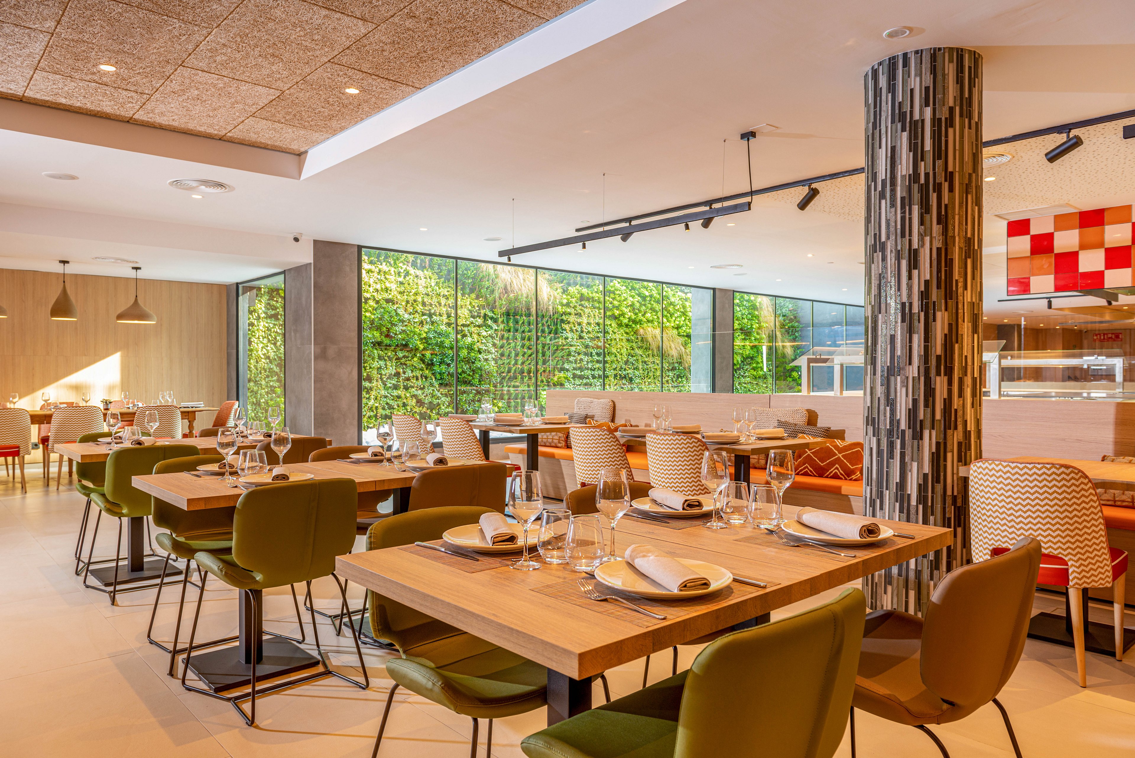 Contemporary restaurant dining area with neatly set wooden tables and green upholstered chairs. The space features large windows with a lush vertical garden wall bringing greenery indoors. Warm pendant lighting and patterned cushions add texture and color to the neutral palette. Glassware, plates, and folded napkins are arranged on each table, ready for guests.