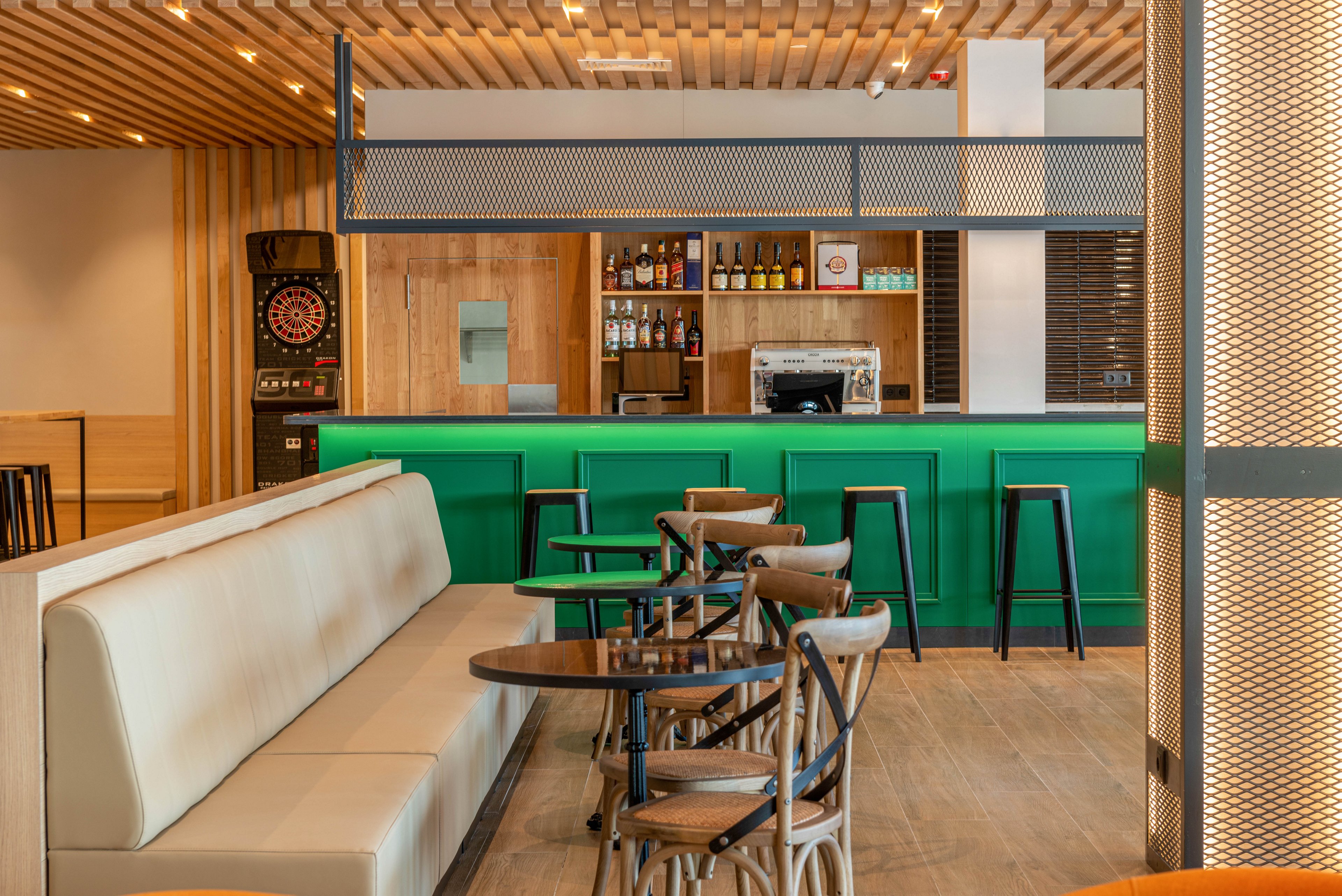 Contemporary cafe bar interior featuring a bright green counter and wooden accents. Empty tables and chairs are arranged beside a long beige upholstered bench. The back bar displays bottles and glassware with a coffee machine and bar stools lined up at the counter. Warm ceiling lights and wood paneling create a cozy, inviting atmosphere, while a dartboard adds a casual entertainment touch.