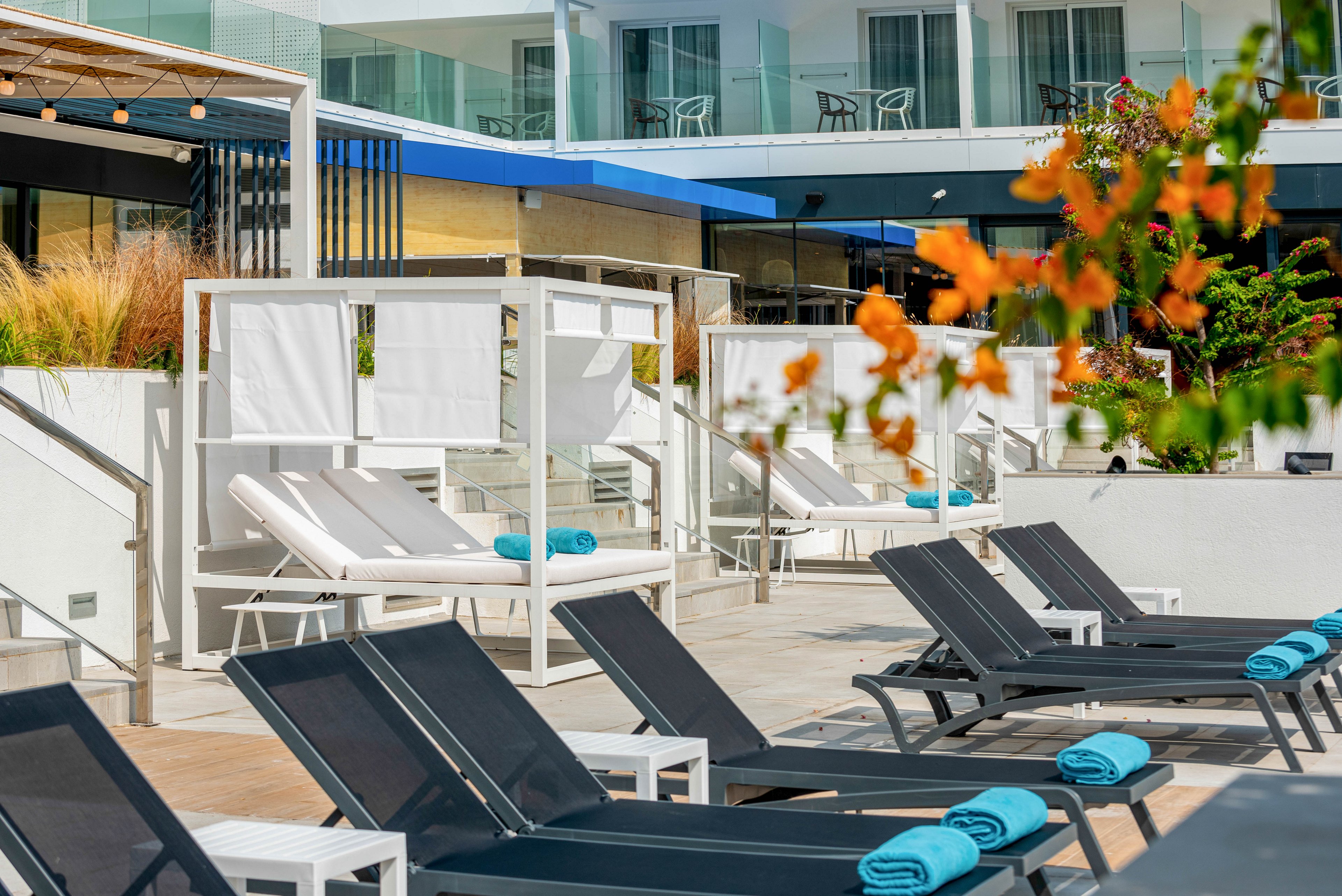 A contemporary hotel poolside area features rows of black and white lounge chairs neatly arranged in the sun. White cabanas with curtains and cushioned daybeds add a touch of luxury to the terrace. Rolled turquoise towels are placed on many of the loungers, providing a bright accent against the neutral tones. The background shows balconies and glass railings of the hotel building, suggesting a relaxing vacation setting.