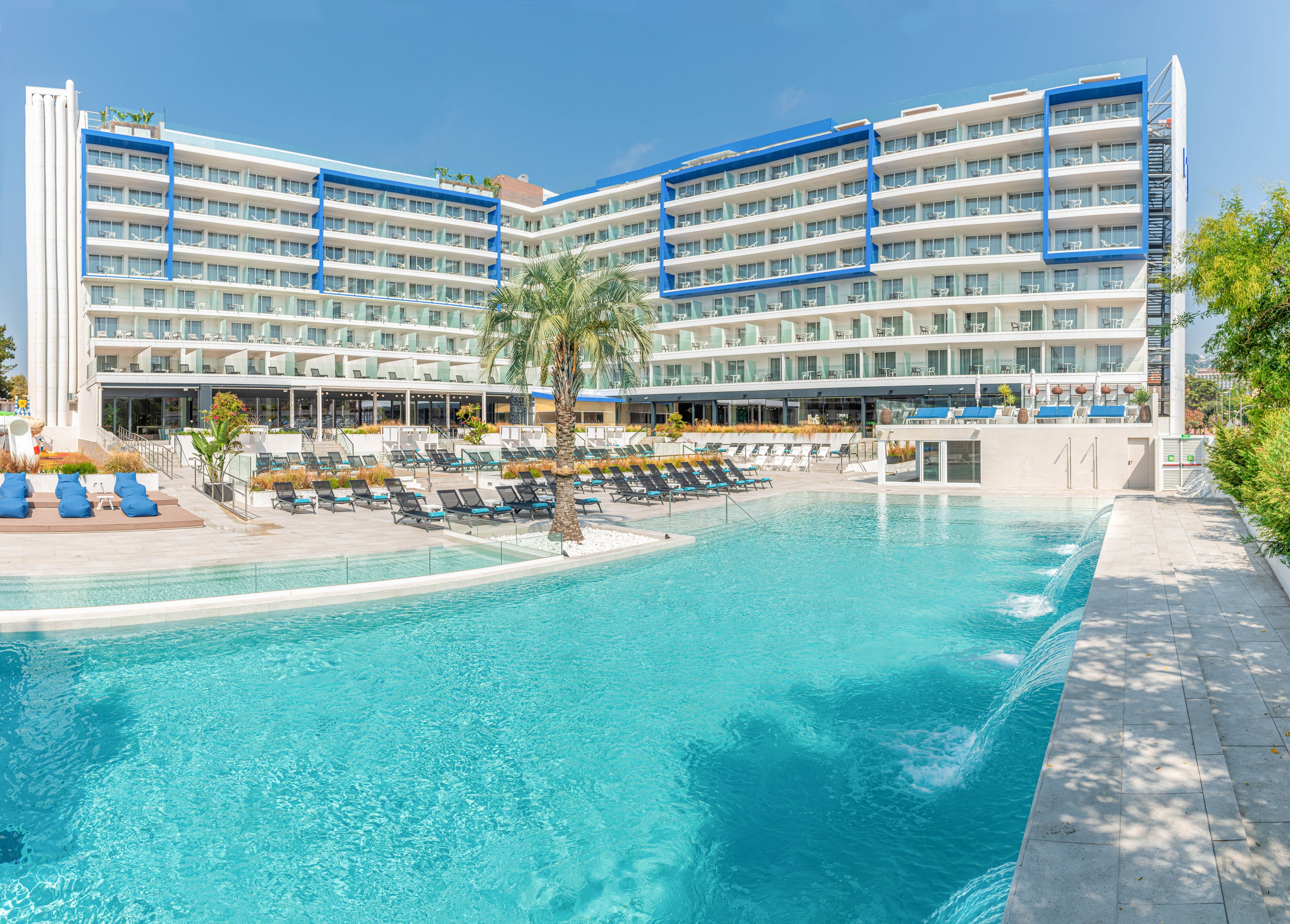 A large contemporary hotel with multiple floors overlooks a wide, turquoise swimming pool. Sun loungers and umbrellas line the poolside terrace, suggesting a relaxing vacation setting. A tall palm tree stands near the center, adding a tropical touch to the scene. The bright blue sky and clean architectural lines create a fresh, modern resort atmosphere.