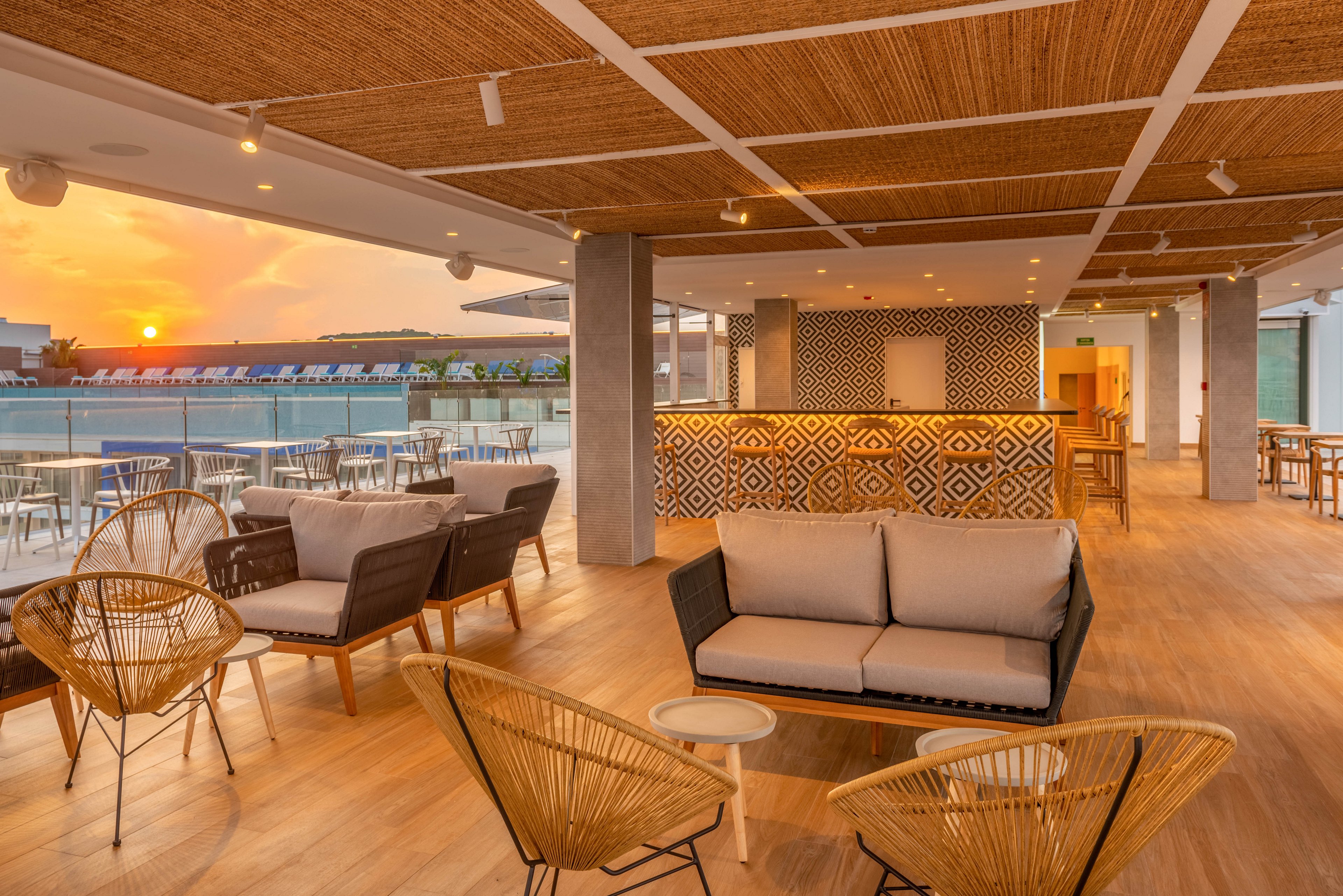Modern rooftop lounge bar with contemporary wicker and cushioned seating is shown during a warm sunset. The space features a patterned bar counter, wooden flooring, and a ceiling with recessed lighting. Outdoor tables and chairs line the terrace, overlooking a pool area and distant horizon. The overall atmosphere feels relaxed and inviting, ideal for evening socializing and leisure.