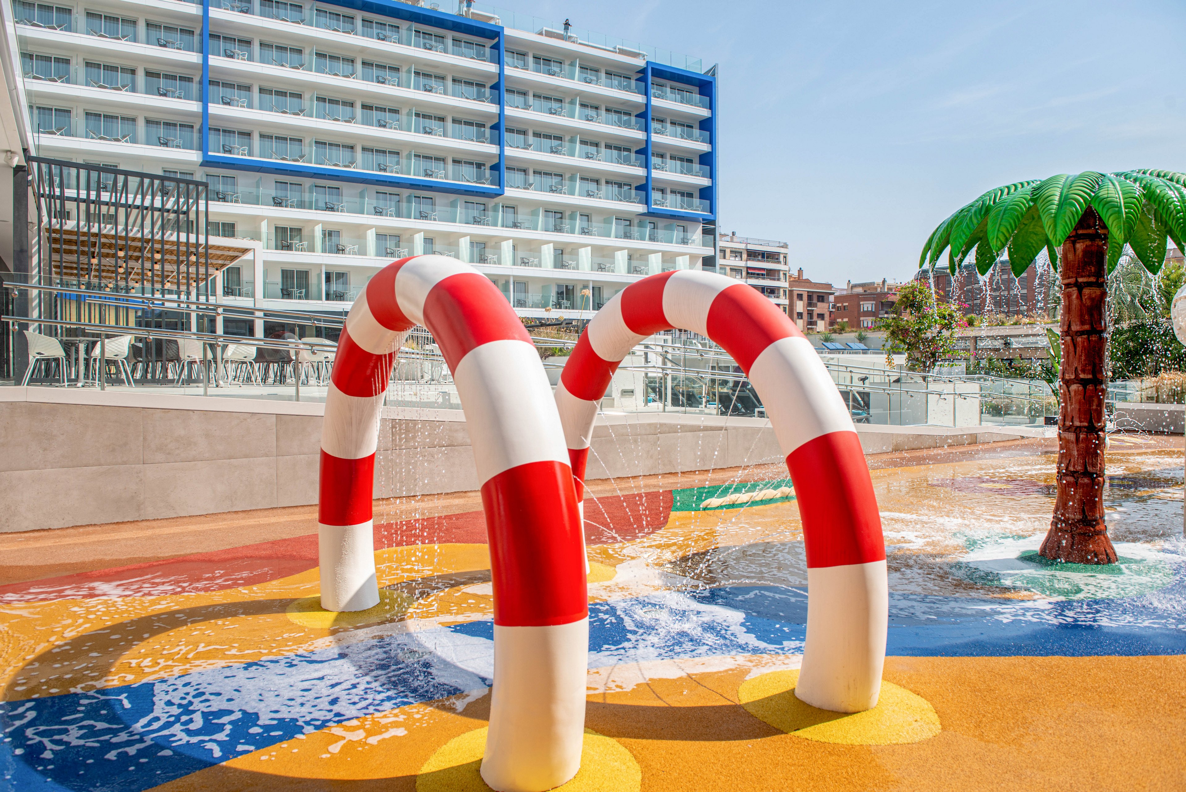 A vibrant splash pad features large red and white striped water arches spraying gentle streams onto the ground. The play area has a colorful, abstract surface with shallow water and playful patterns. A tall palm tree-shaped fountain stands to the side, adding to the tropical theme. In the background, a modern multi-story hotel with balconies overlooks the water play area under a clear blue sky.