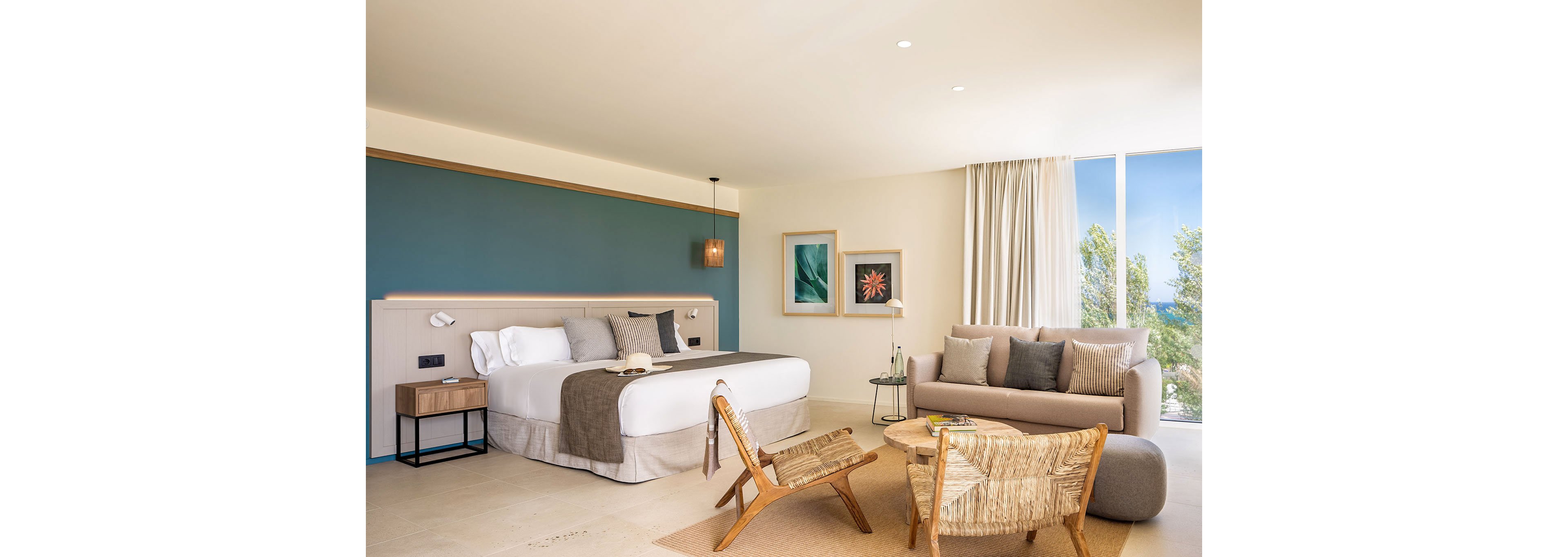 Bright contemporary hotel room featuring a large bed with neutral linens and accent pillows. A cozy seating area with a sofa, woven chairs, and a small round table creates a relaxed lounge space. Natural light floods in through floor-to-ceiling windows, revealing greenery outside. Minimalist decor, soft earth tones, and framed artwork on the wall contribute to a calm, inviting atmosphere.