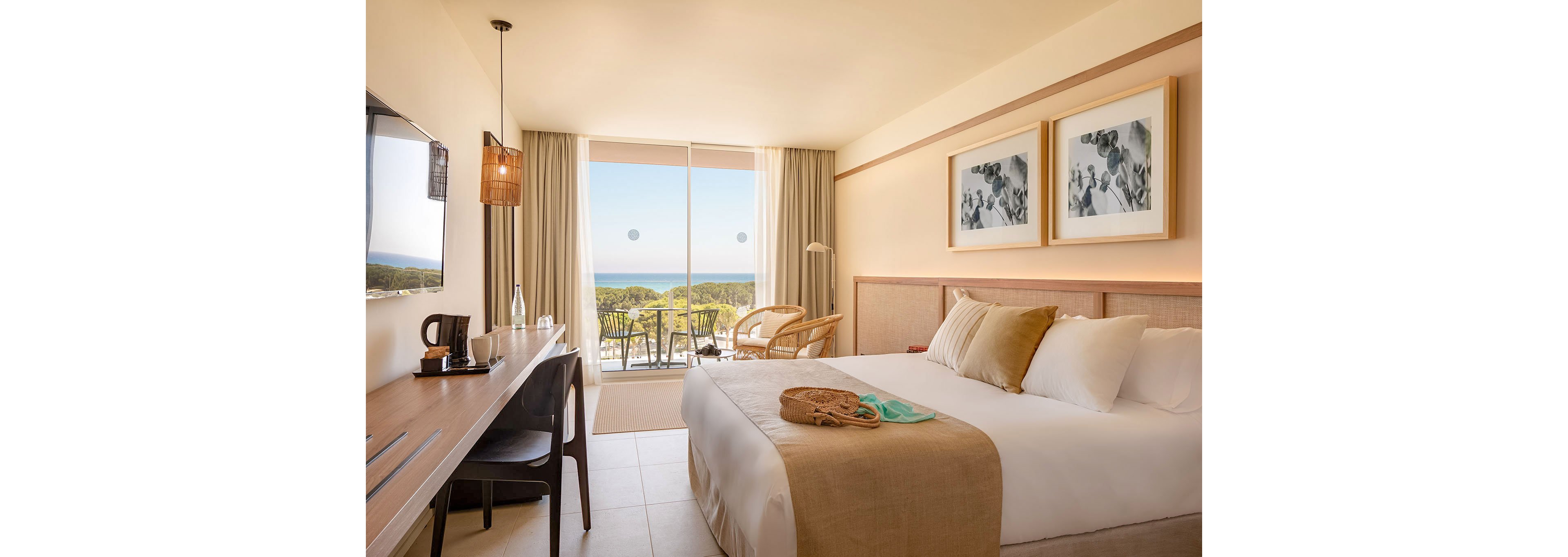A modern hotel room features a neatly made double bed with neutral-toned pillows and throw. A desk with a chair, lamp, and kettle sits along one wall, facing a flat-screen TV. Large sliding glass doors open to a balcony with outdoor seating and a scenic view of trees and the distant sea. Soft beige and white colors, natural light, and simple framed artwork create a calm, inviting atmosphere.