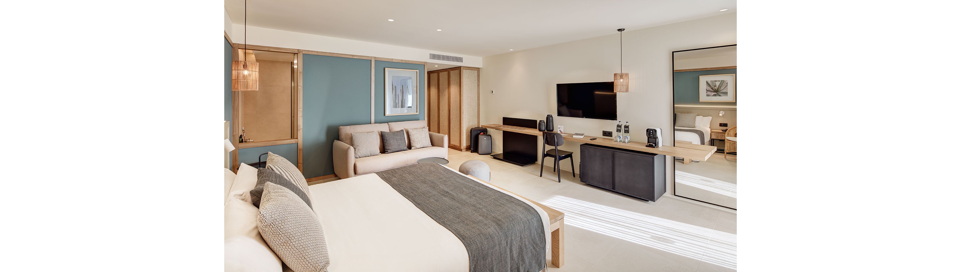 Spacious contemporary hotel room featuring a large bed with a gray throw and light bedding. A comfortable sofa sits against a teal accent wall beside a framed artwork. Opposite the bed, a sleek desk and chair face a wall-mounted flat-screen TV, with coffee machines and bottles arranged neatly. Warm neutral tones, minimalist furniture, and soft natural light create a calm, inviting atmosphere.