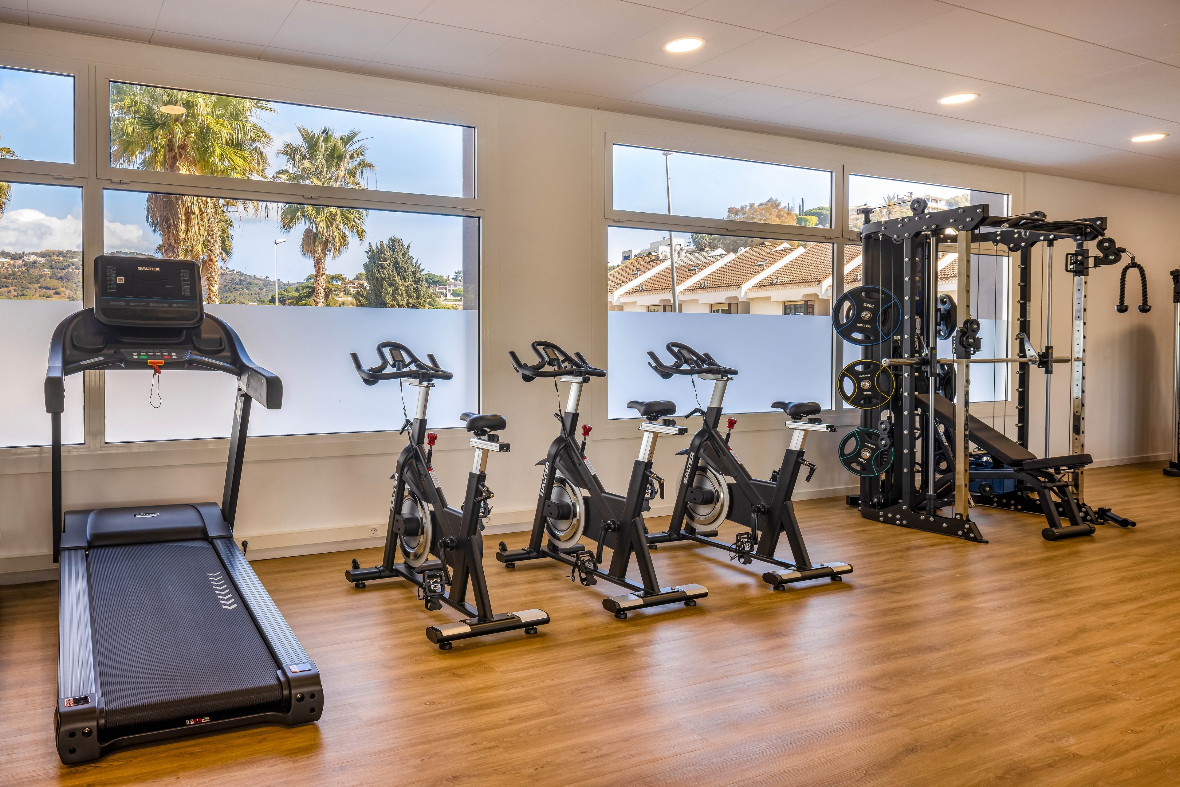 A bright, modern fitness room features a row of stationary bikes, a treadmill, and a large multifunction strength-training machine. The gym has wood-style flooring and large windows that let in natural light. Palm trees and a sunny outdoor landscape are visible through the windows, creating an inviting atmosphere. The space appears clean, organized, and ready for individual workouts or small group exercise sessions.