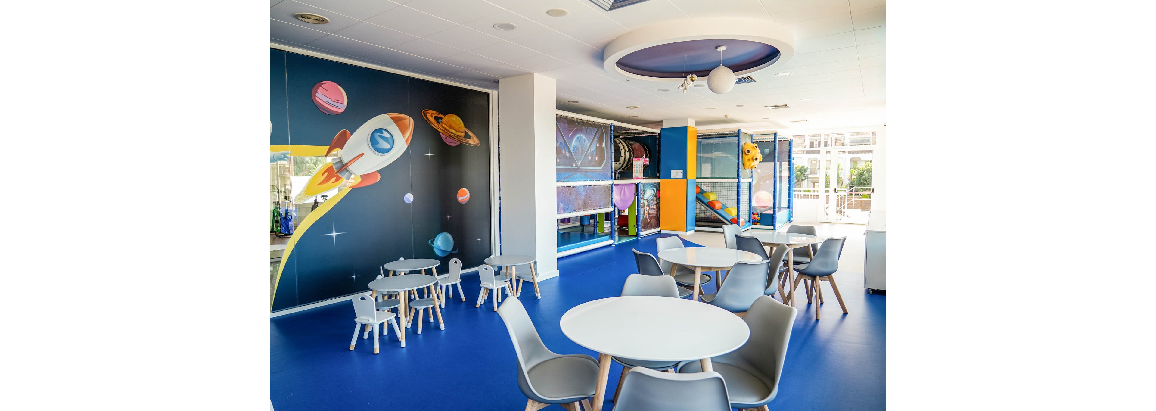 Bright indoor children's playroom decorated with a space theme. Small round tables and child-sized chairs are arranged on a blue floor. The walls feature colorful illustrations of rockets and planets, adding a playful atmosphere. Large windows and ceiling lights provide ample natural and artificial light throughout the room.
