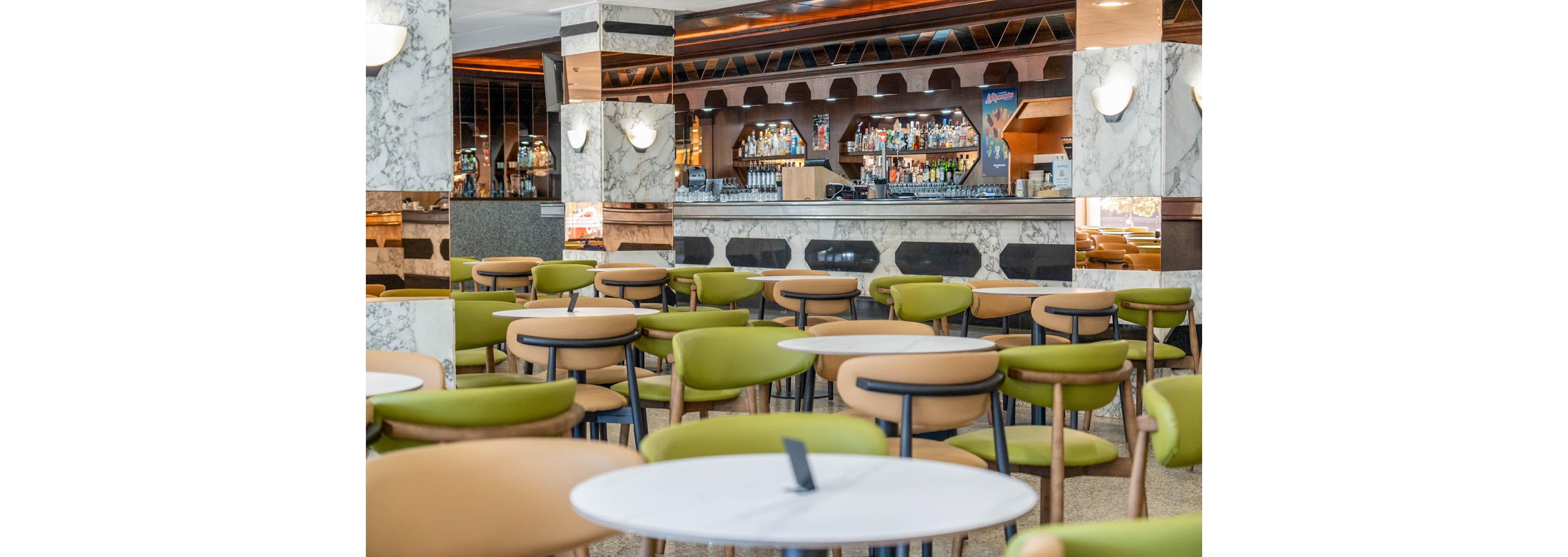 A contemporary bar area features rows of round tables surrounded by green and tan cushioned chairs. The space appears empty, with no people present, emphasizing the clean and orderly arrangement of the furniture. In the background, a well-stocked bar with bottles, glassware, and decorative arches is framed by marble columns and warm wood accents. Soft wall lighting and reflective surfaces contribute to a bright, inviting atmosphere suitable for socializing or relaxing.