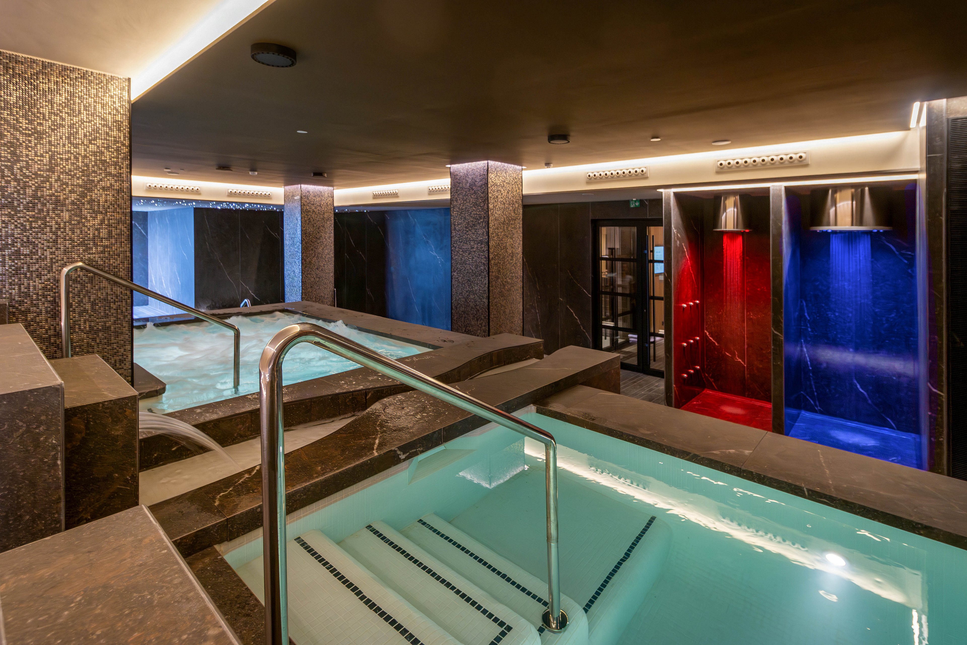 A contemporary indoor spa features multiple turquoise plunge pools with stainless steel handrails. The space is finished with dark stone walls and warm recessed ceiling lighting. In the background, individual shower or steam areas are lit with dramatic red and blue accent lights. The overall atmosphere is calm, luxurious, and designed for relaxation and hydrotherapy.