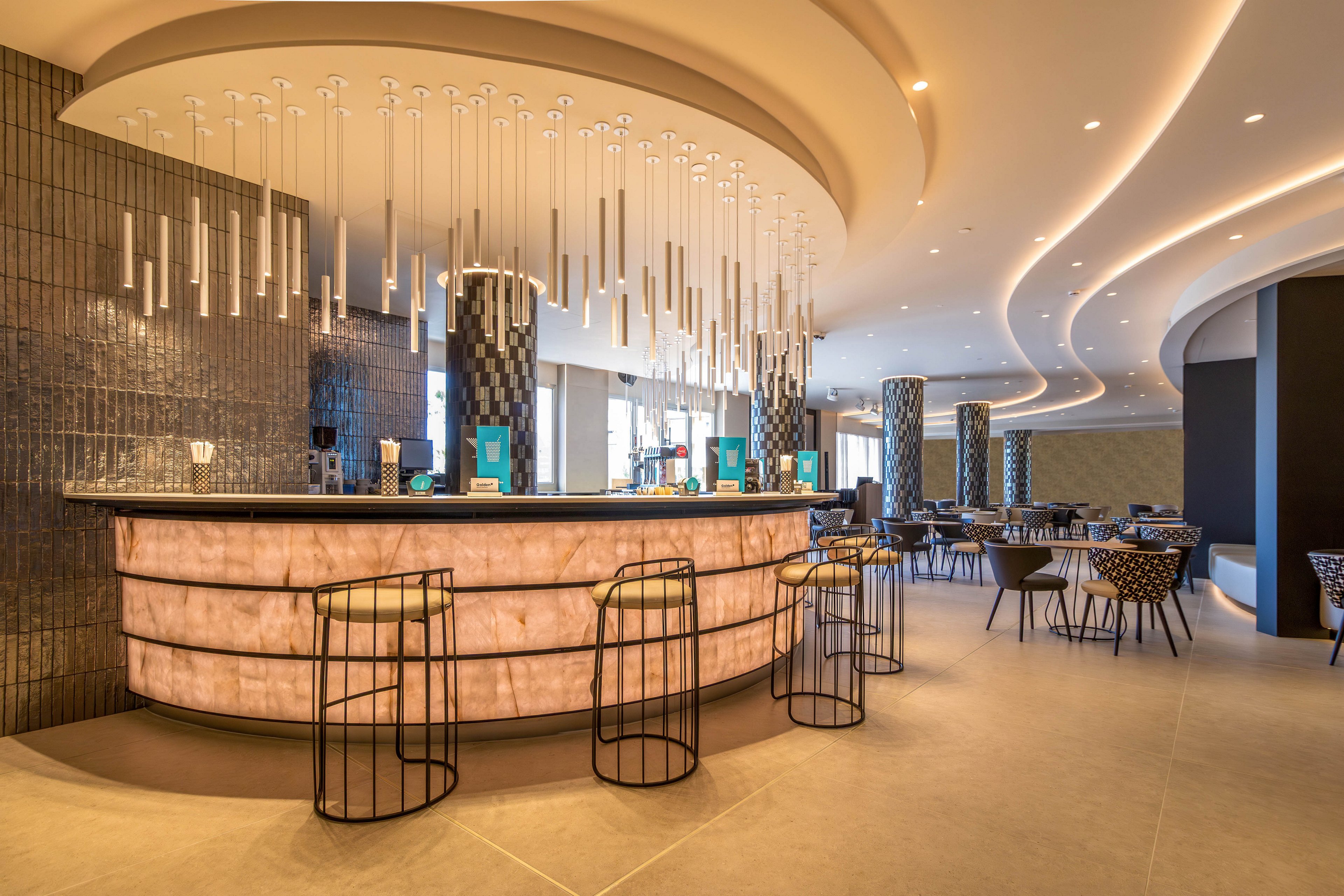 A bartender stands behind a curved, illuminated bar counter in a contemporary hotel lounge. The space features sleek bar stools, patterned dining chairs, and small round tables arranged around the room. Long pendant lights hang from the ceiling, complementing the flowing, curved ceiling design. Warm lighting and neutral tones create an inviting, upscale atmosphere suitable for socializing or relaxing.